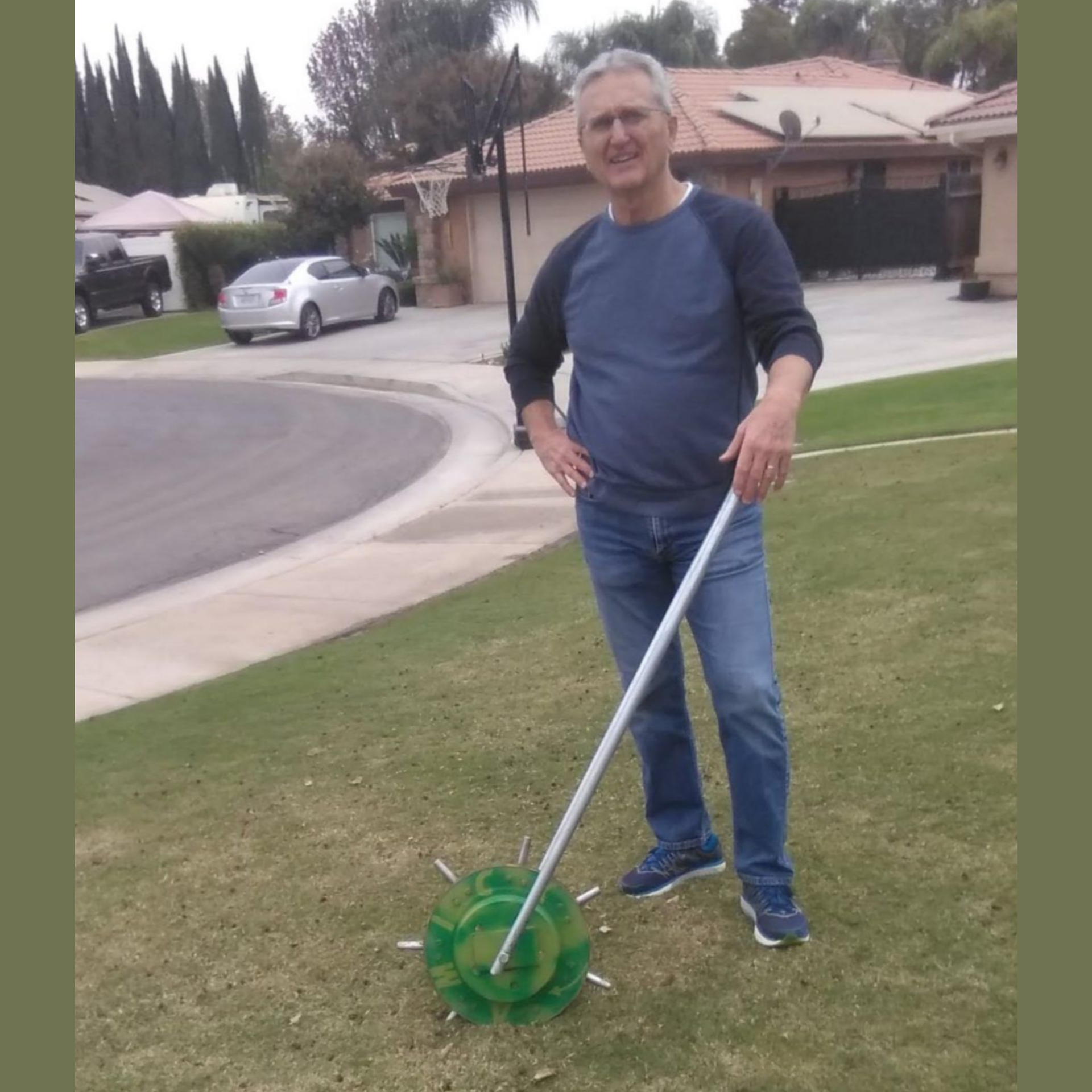 A person stands on a suburban lawn holding a long-handled tool with a green, multi-pronged metal attachment at the base.