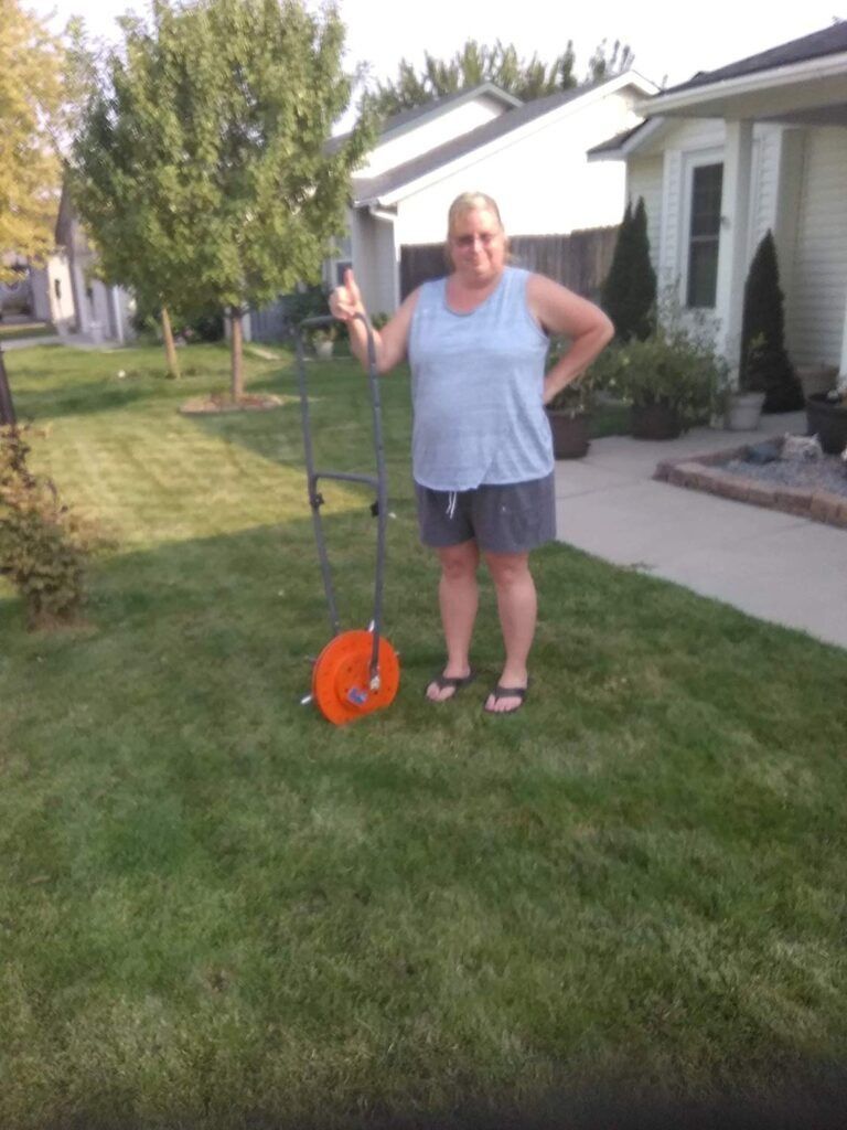 A person stands in a grassy yard holding a large orange measuring wheel tool next to a house.