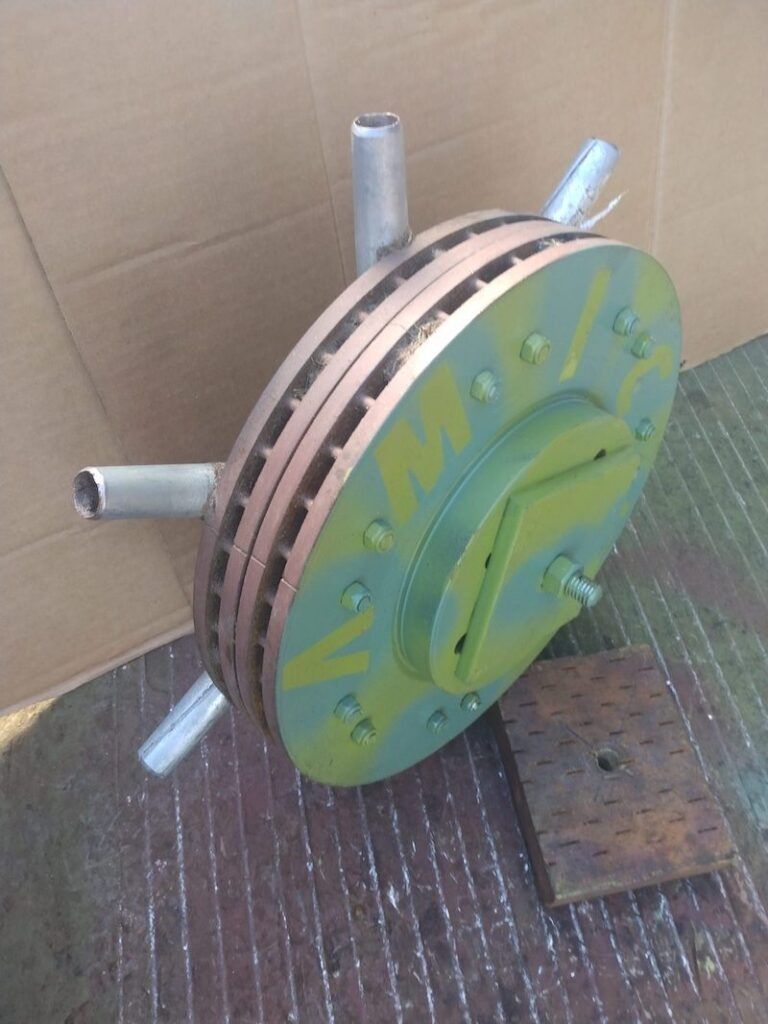 A vented, yellow-painted circular mechanical component with radiating metal tubes, sitting on a wooden block.