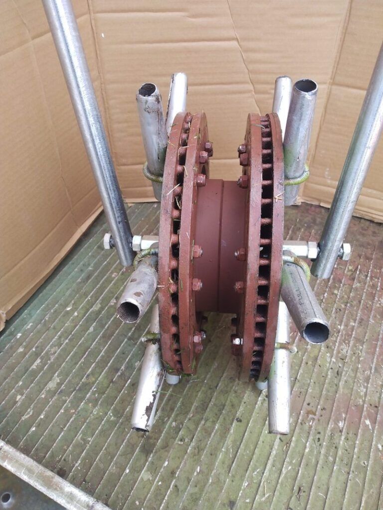 A brown, dual-disk industrial component held by metal pipes against a cardboard backdrop.