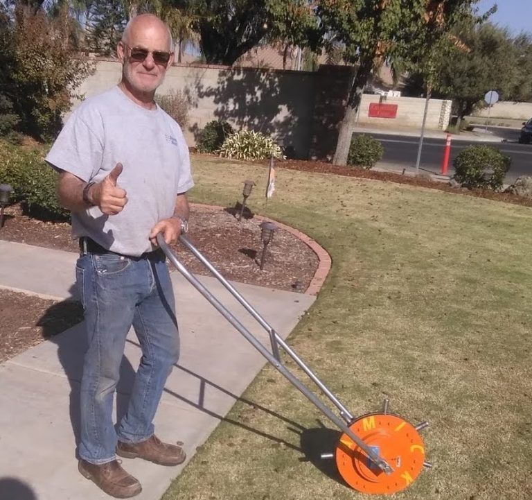 A person stands on a sidewalk, giving a thumbs up while holding a rolling measuring wheel on a grassy lawn.