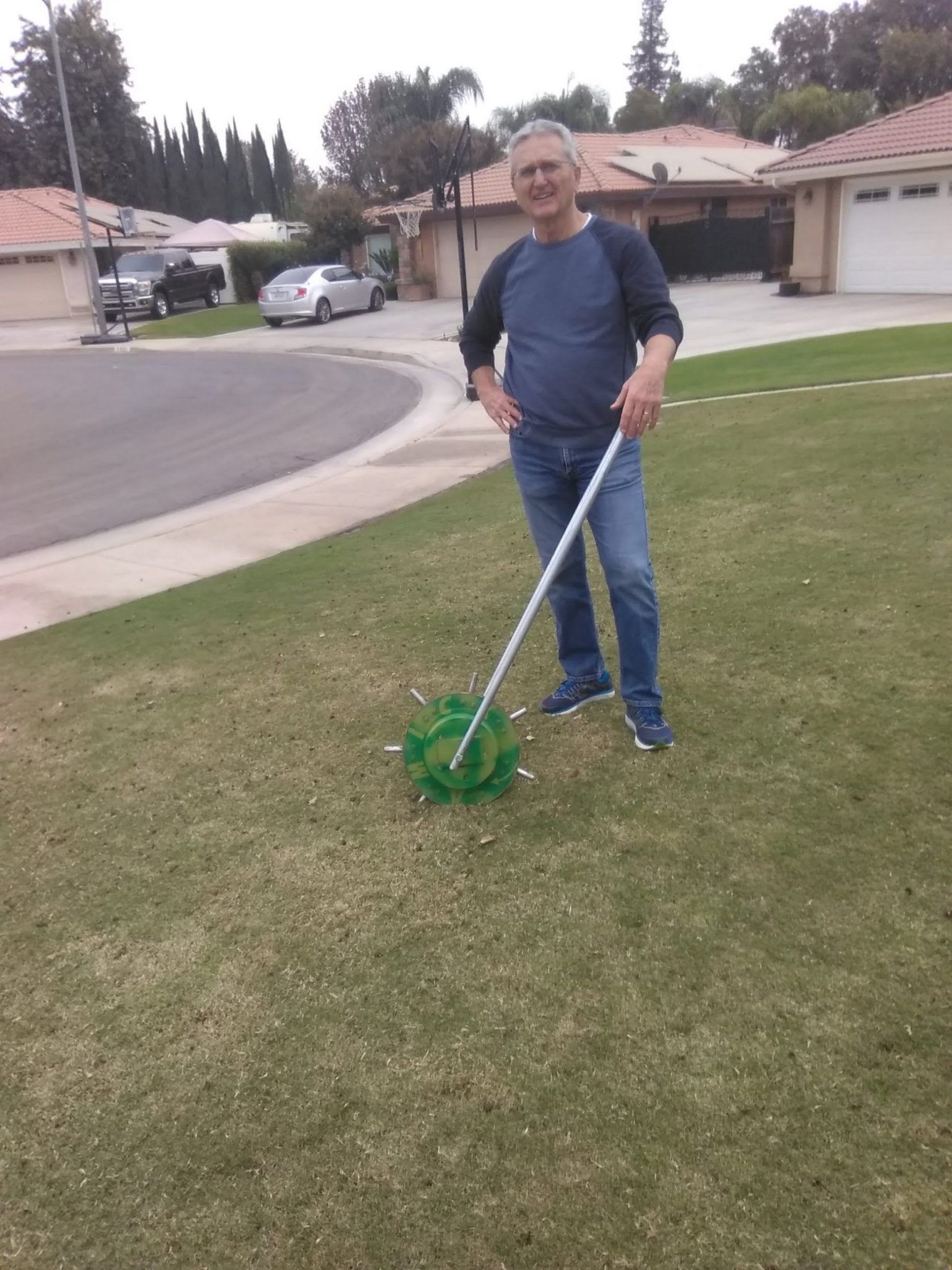A person stands on a suburban lawn holding a long-handled tool with a green, multi-pronged metal attachment at the base.
