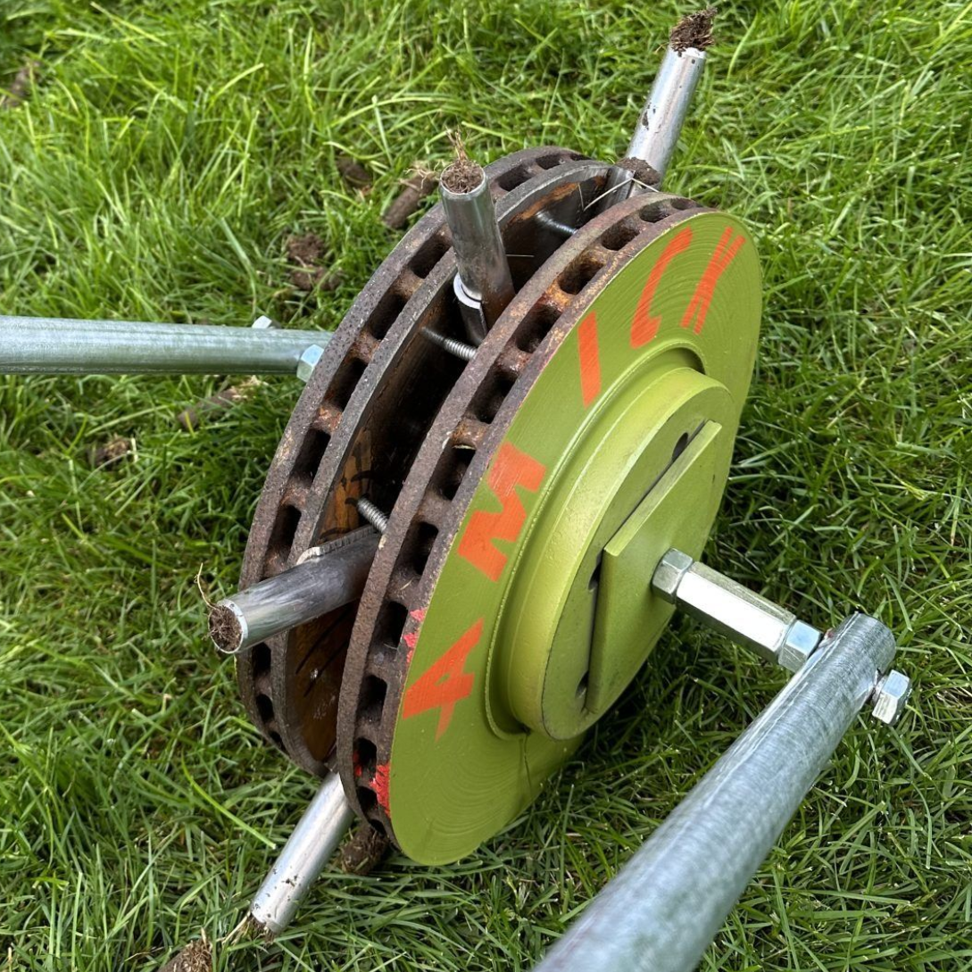 A green-painted metal brake disc assembly with protruding metal rods attached to a frame, resting on green grass.