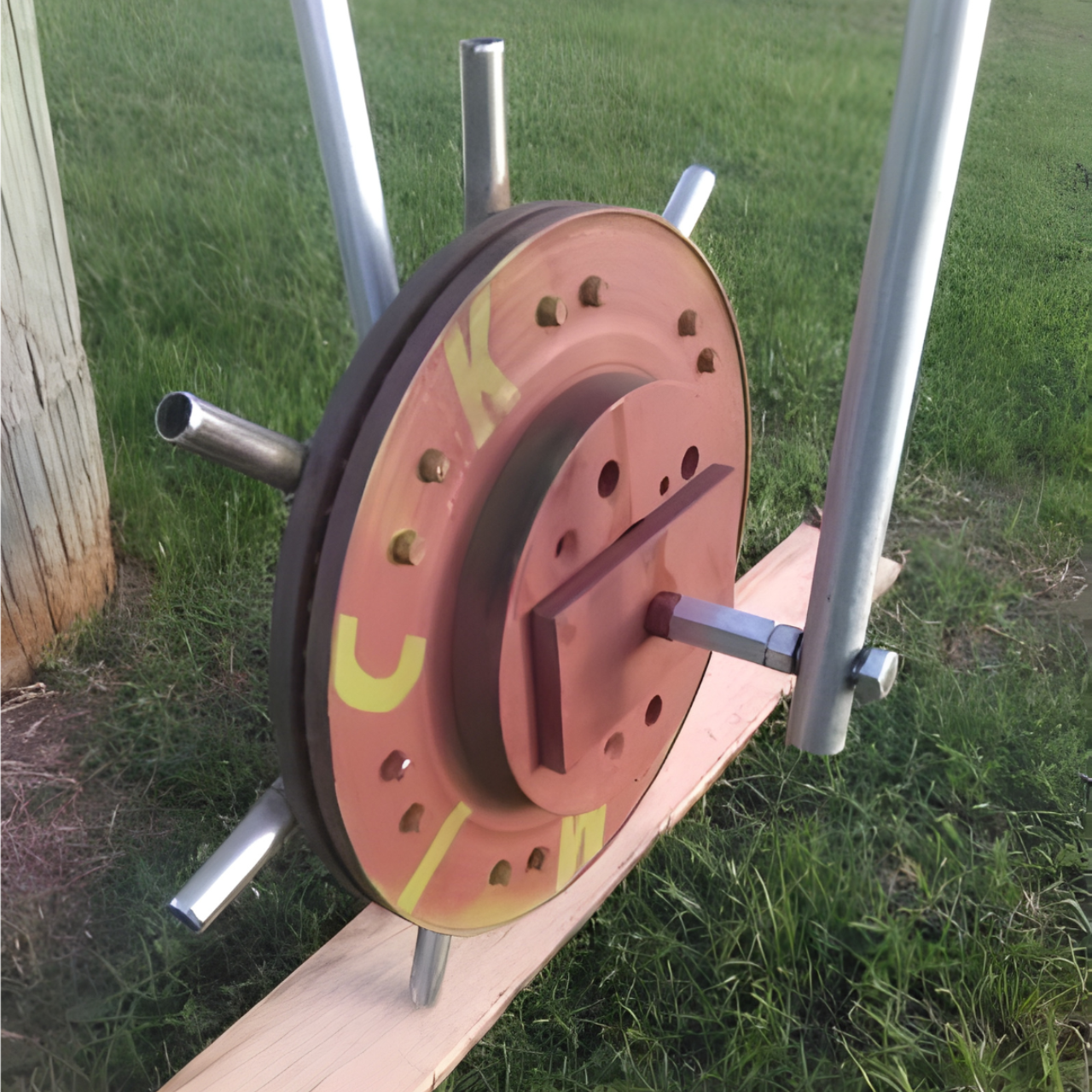 A handmade rotating sculpture featuring a painted metal wheel with protruding steel tubes, mounted on a wood base in grass.