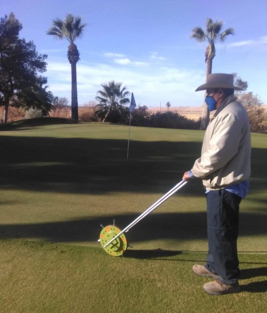 A person wearing a hat and mask uses a golf stimpmeter to measure the speed of the grass on a putting green.