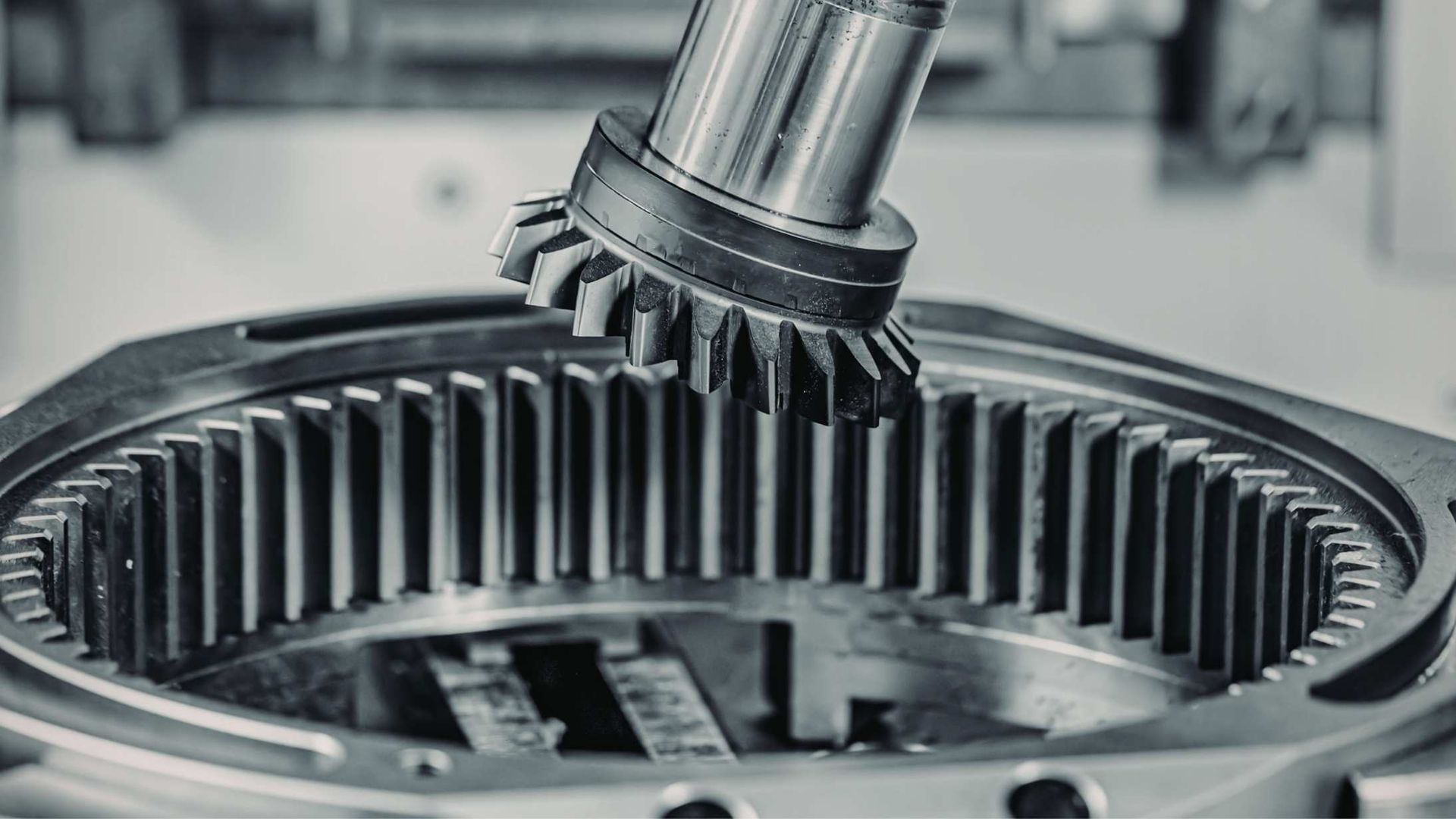 A metal pinion gear is positioned above a large, stationary internal ring gear in an industrial manufacturing setting.