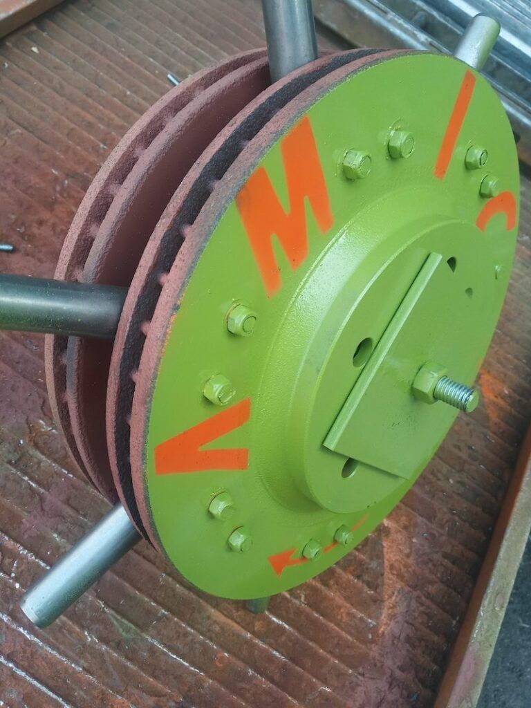 A green metal disc assembly with orange letters, joined to brake rotors and metal rods, sitting on a ribbed surface.