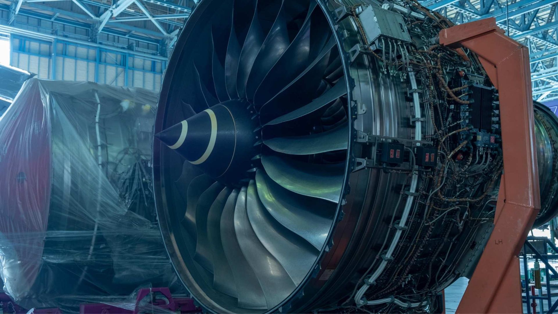 A large jet engine with curved silver blades stands on a red support stand inside a maintenance facility.
