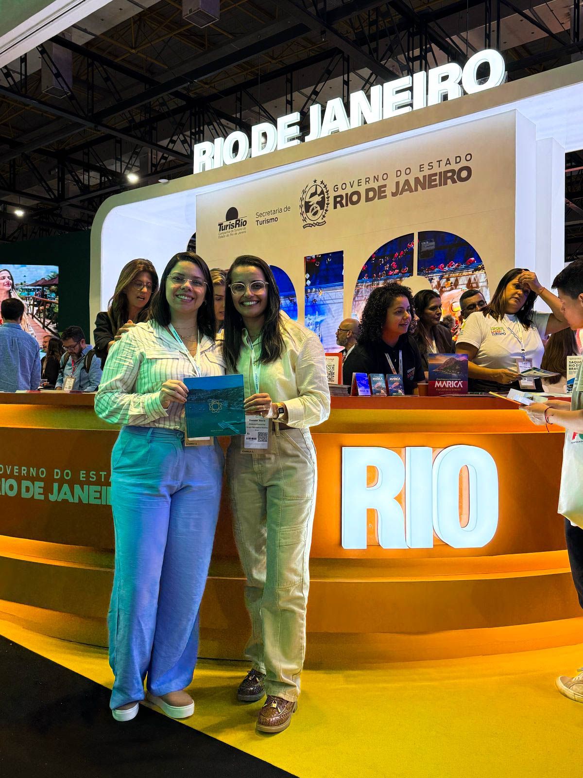 Duas mulheres estão de pé, uma ao lado da outra, em frente a uma placa do Rio. 