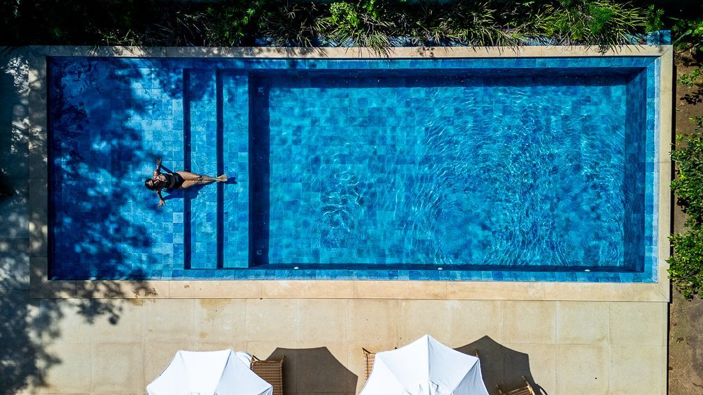 Woman floating in a blue pool, viewed from above, two white umbrellas below.