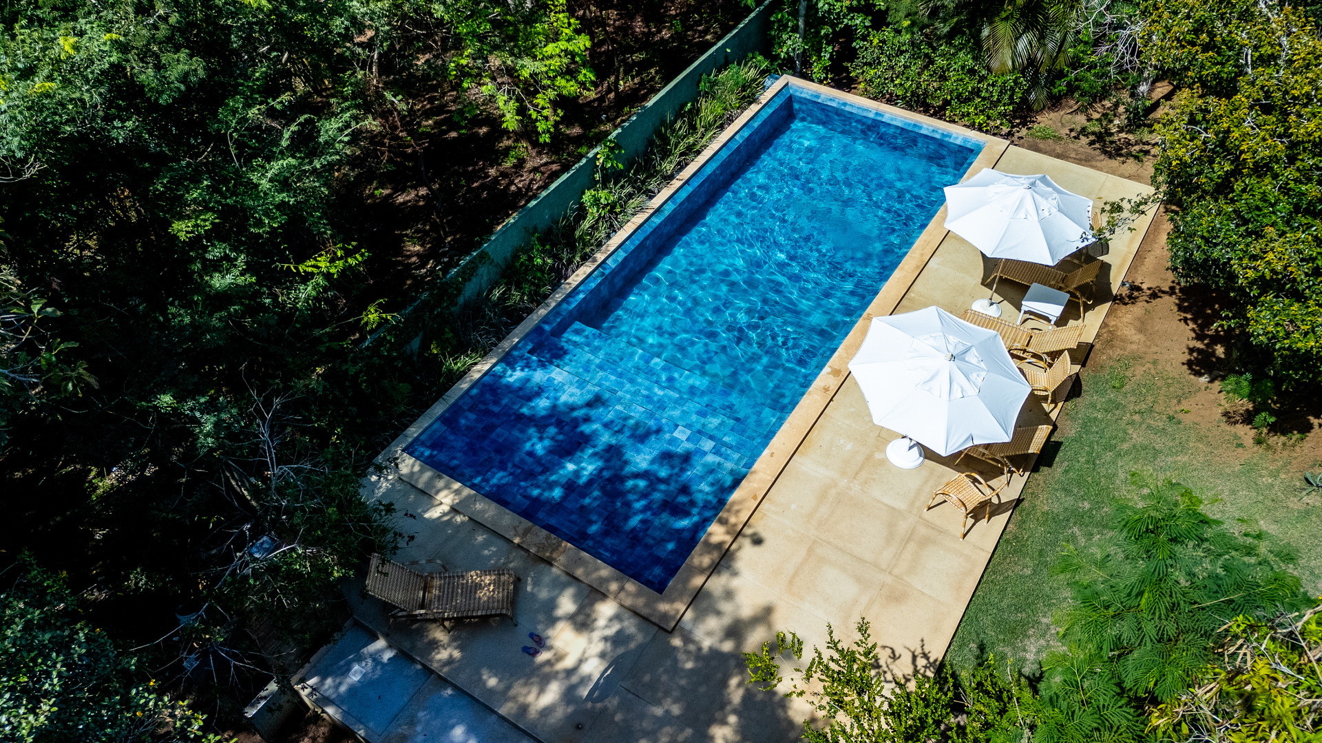 Uma vista aérea de uma grande piscina com guarda-sóis e cadeiras cercada por árvores.