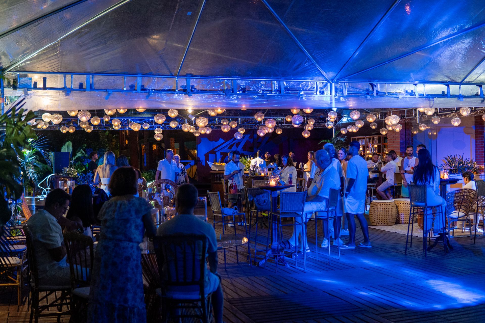 People at an outdoor event under a lit canopy. Blue lighting illuminates the space, with string lights overhead.