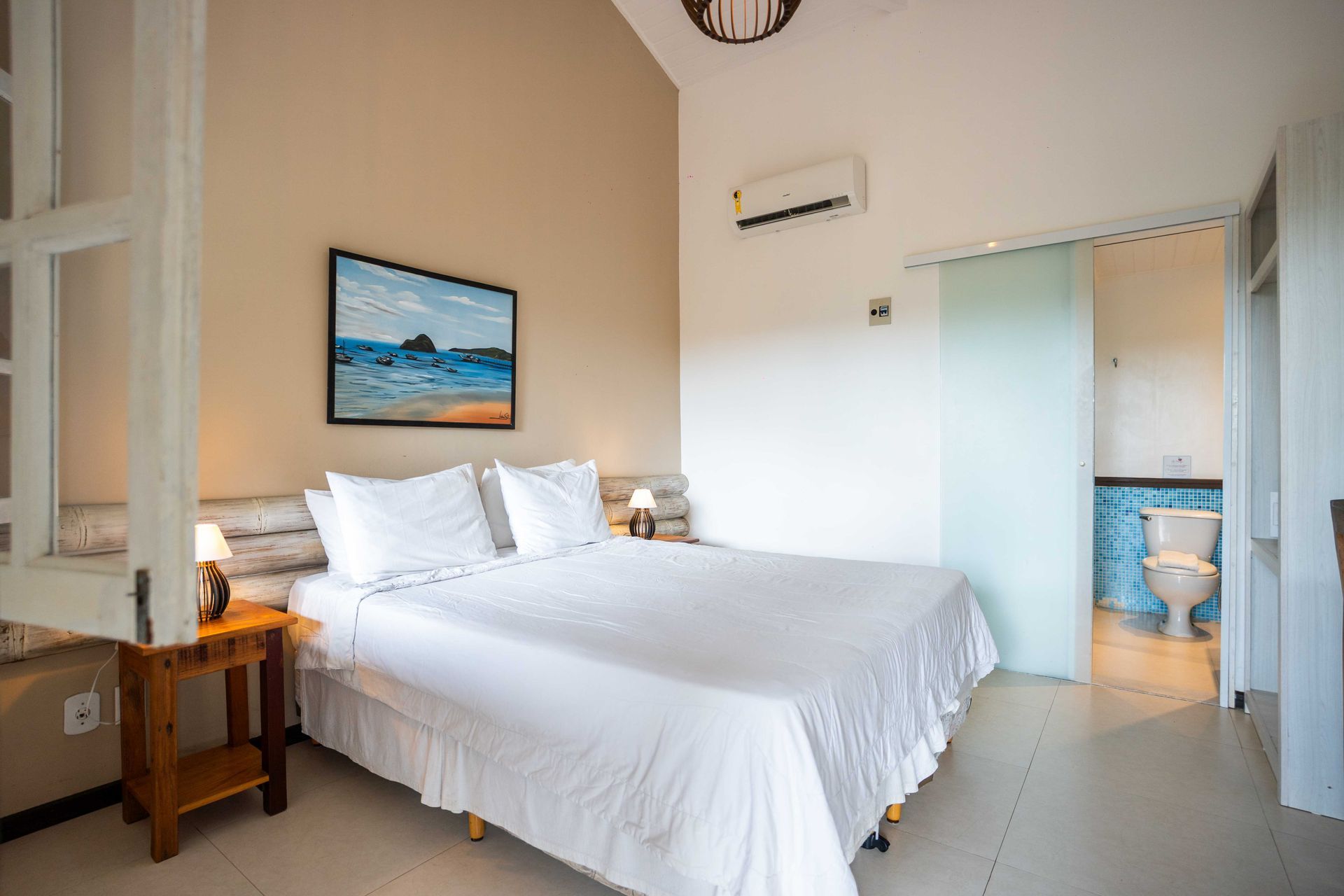Um quarto com uma cama king size, móvel ao lado da cama, um quadro acima da cama, ar-condicionado e uma vista para o banheiro.
