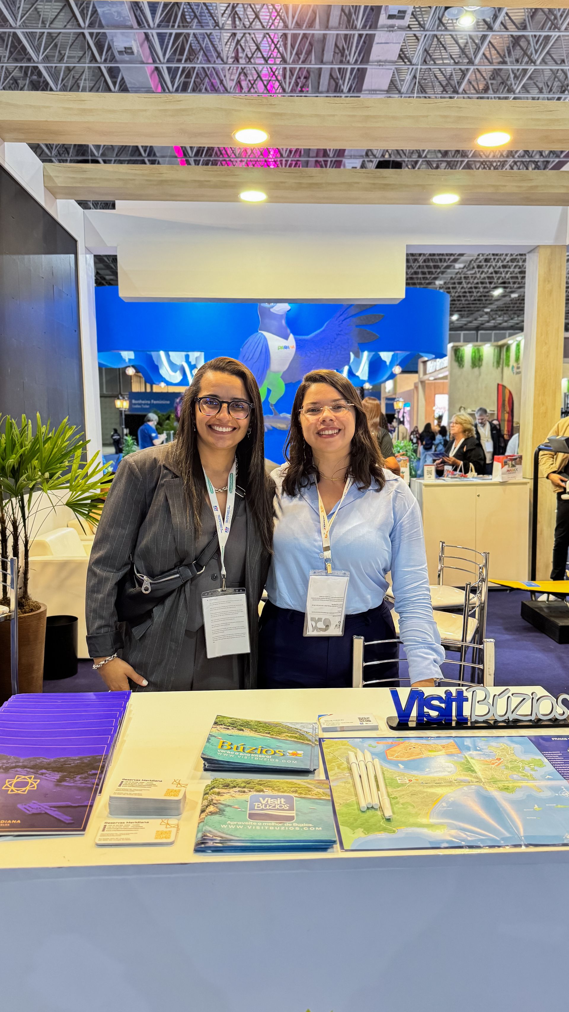 Duas mulheres em um estande de uma feira de turismo. Uma de blazer, a outra de camisa de botões, ambas sorrindo. O estande exibe folhetos de viagens.