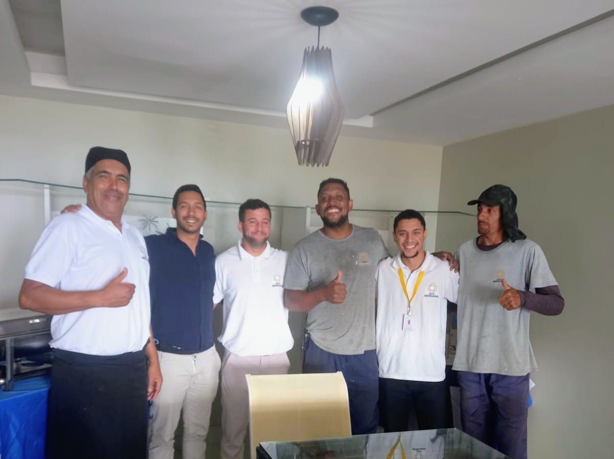 Group of six people giving thumbs up indoors. Men are diverse in appearance, wearing various shirts and pants.
