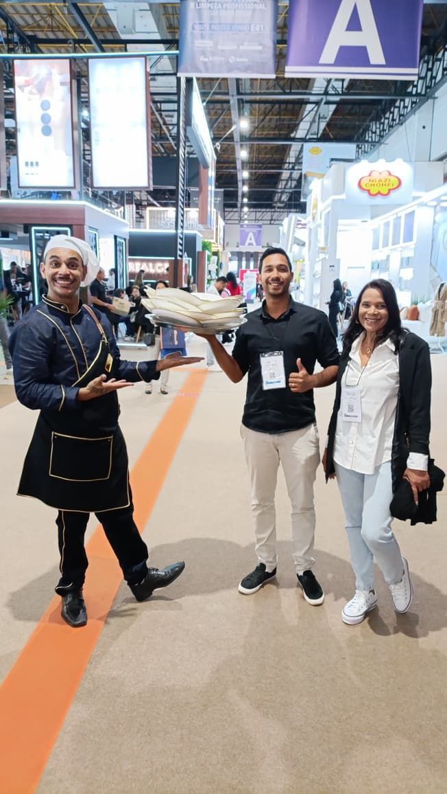 A man in chef attire offers food to a smiling couple at a trade show.