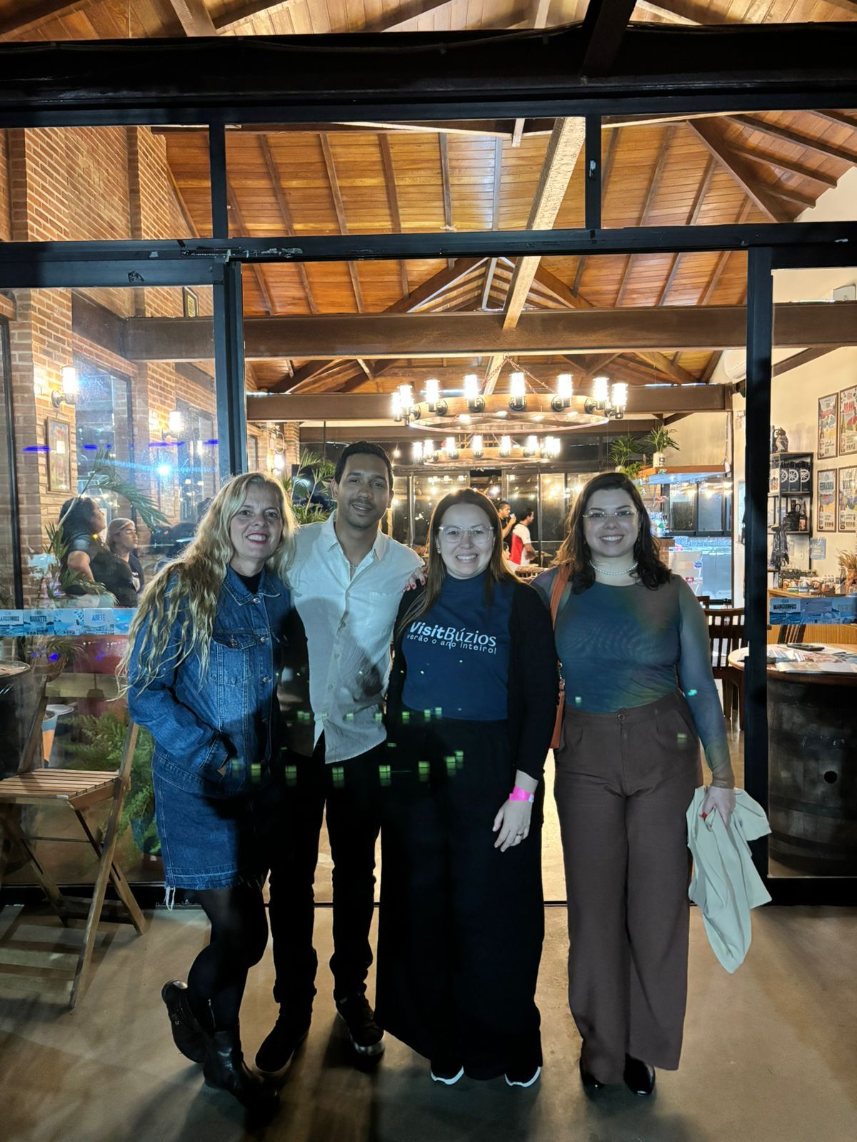 Four people pose in front of a restaurant entrance at night. People are smiling and holding green bottles.