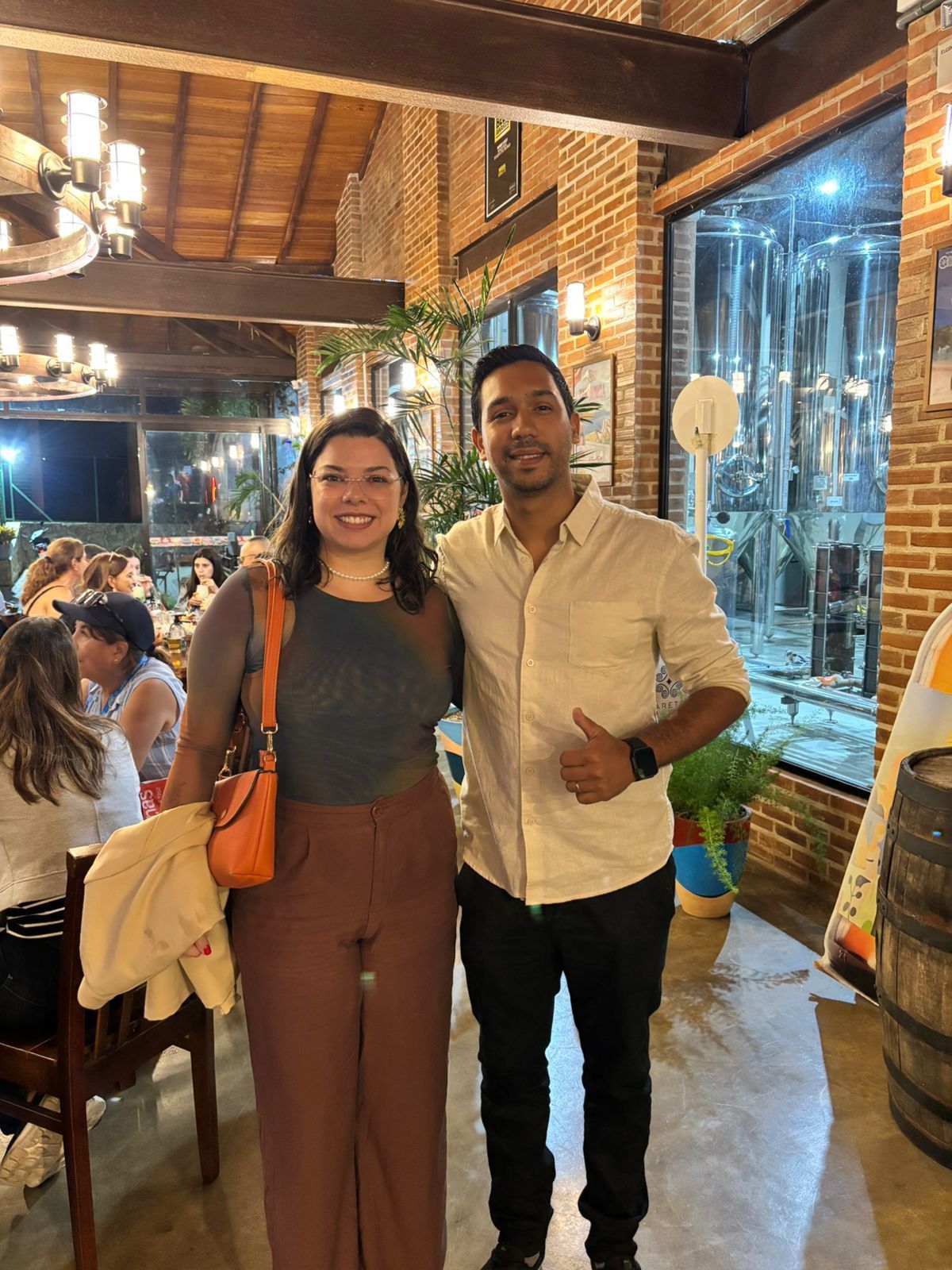 A smiling couple poses for a photo in a restaurant. The woman wears brown pants, a gray top, and a shoulder bag. The man wears a light shirt and black pants.