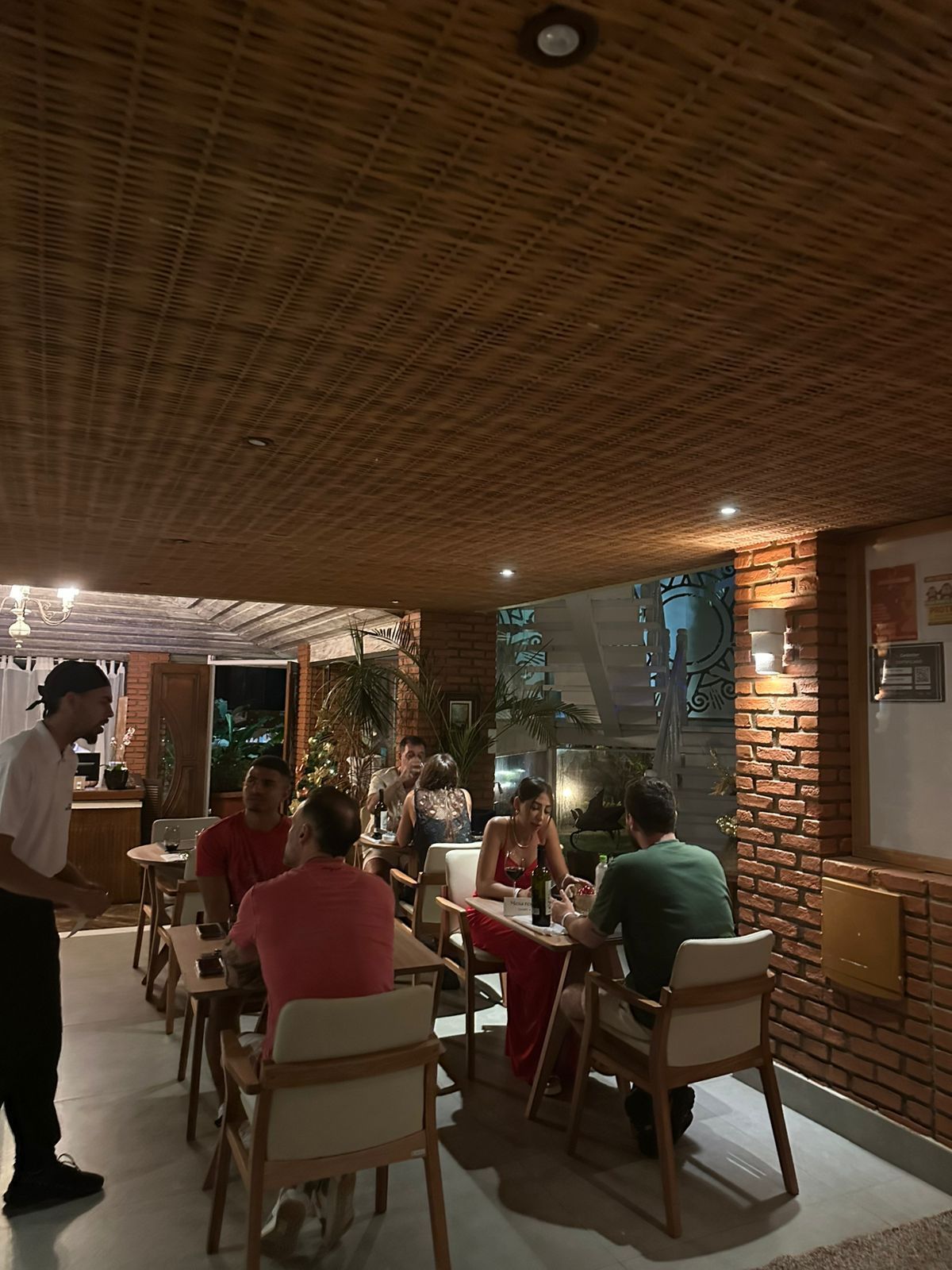 Interior de um restaurante com clientes sentados às mesas. Um garçom está próximo.
