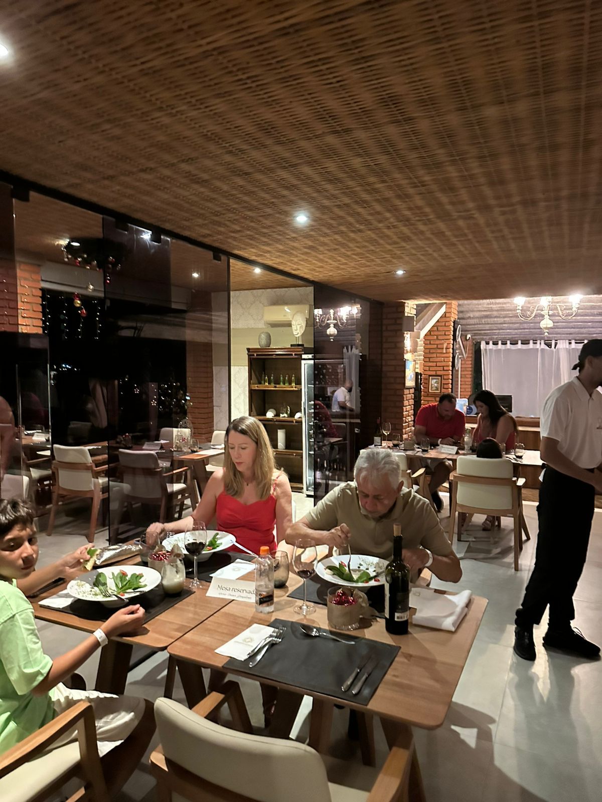 Pessoas jantando em um restaurante com mesas de madeira e um garçom.