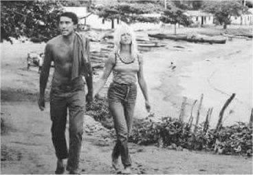 Homem com uma toalha e mulher de calça jeans caminhando por uma trilha na praia; árvores e prédios ao fundo.