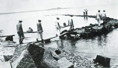 Workers building a stone causeway across water, using planks to walk and carry materials.
