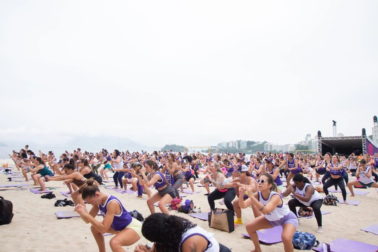 Grupo de pessoas fazendo agachamentos em uma praia, sob um céu nublado.