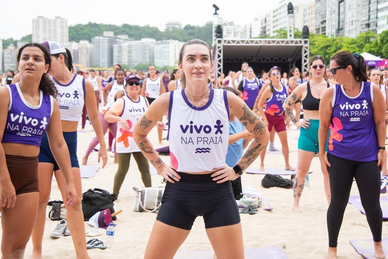 Group of people exercising on a beach; main subject with tattoos, wearing white tank, black shorts, smiling.