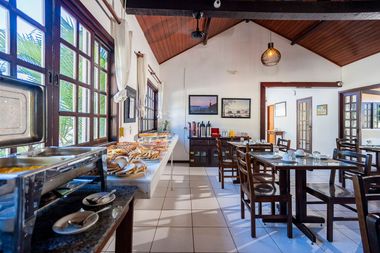 Área de café da manhã estilo buffet com exposição de alimentos, mesas e cadeiras. Grandes janelas à esquerda proporcionam iluminação natural.