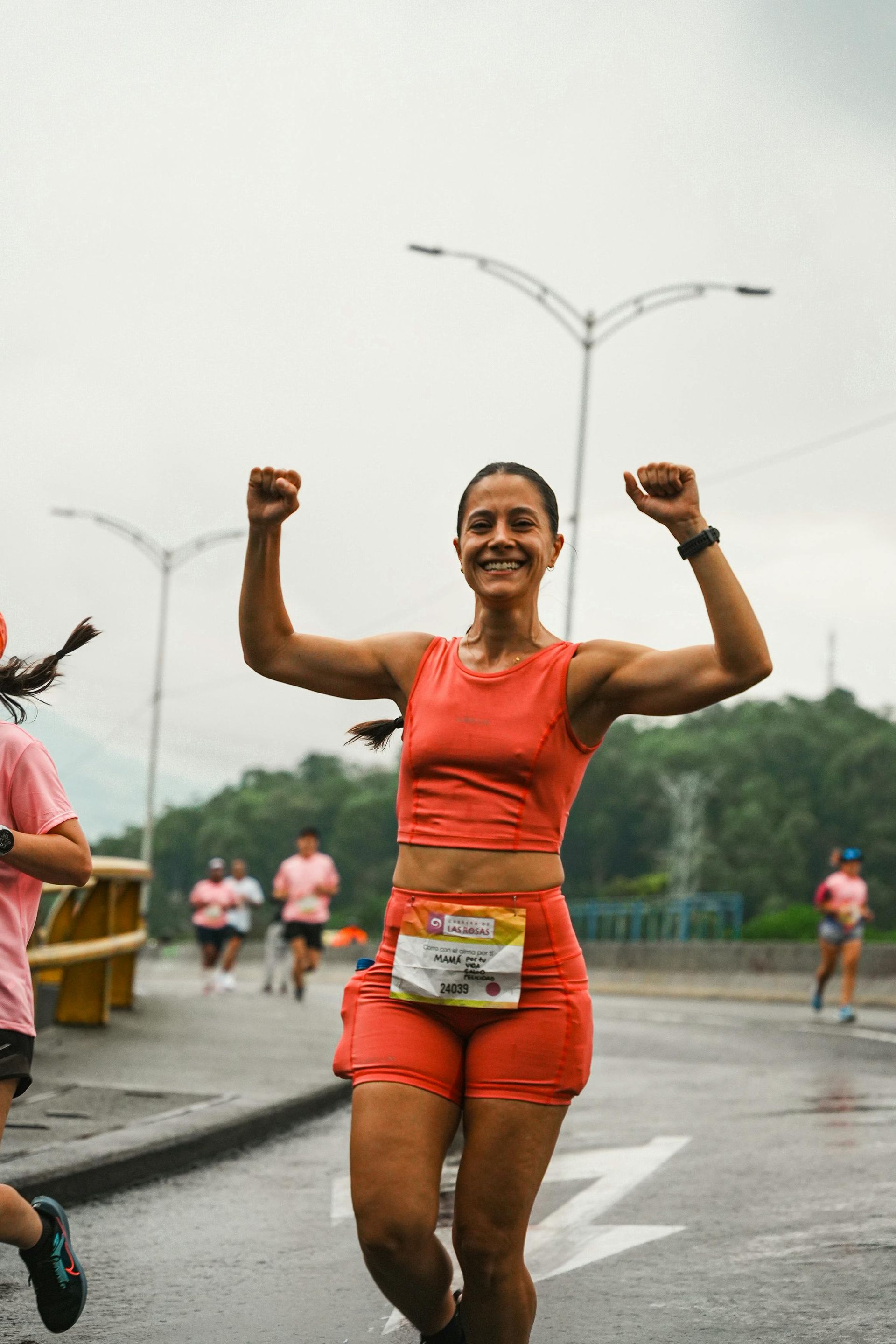 A runner in an orange top and shorts smiles with arms raised in celebration during a race on a paved road.