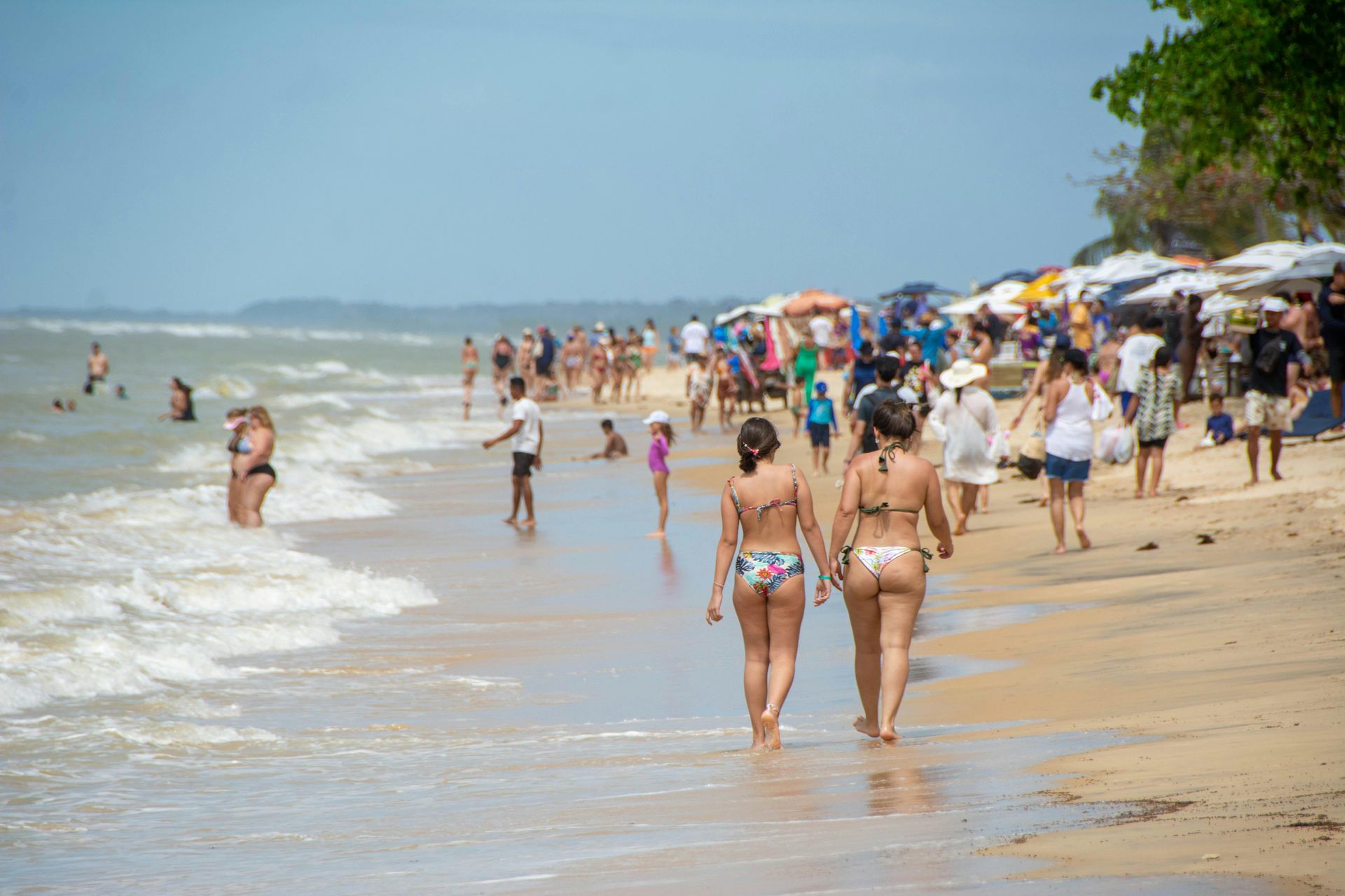 People in swimwear walk and relax along a sunny, crowded beach with waves gently washing onto the sand.