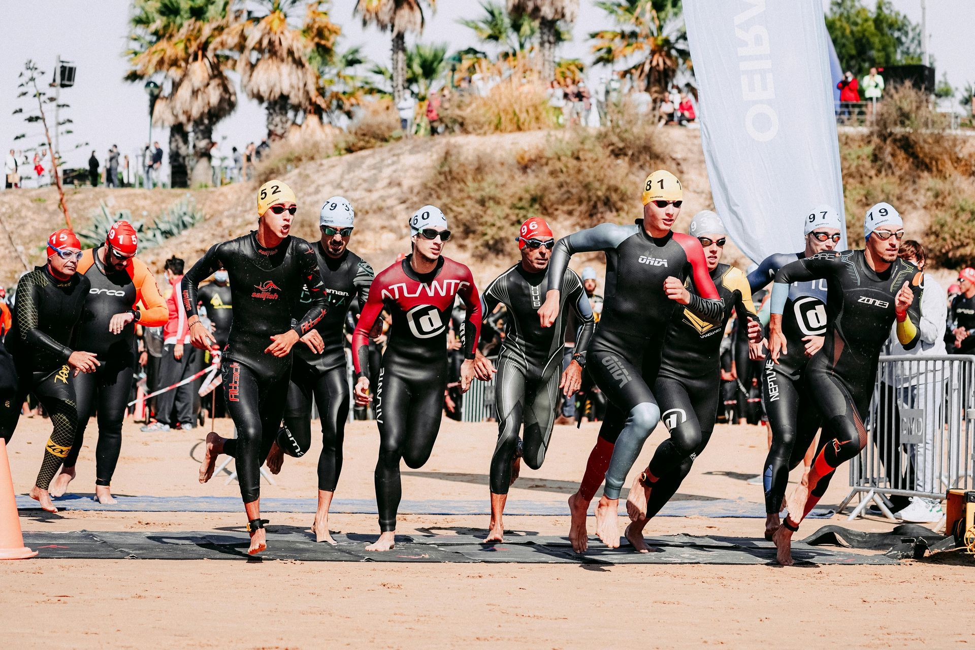 Triatletas com trajes de neoprene e toucas de natação começam a correr em uma praia de areia a partir de um tapete, com uma colina ladeada por palmeiras ao fundo.