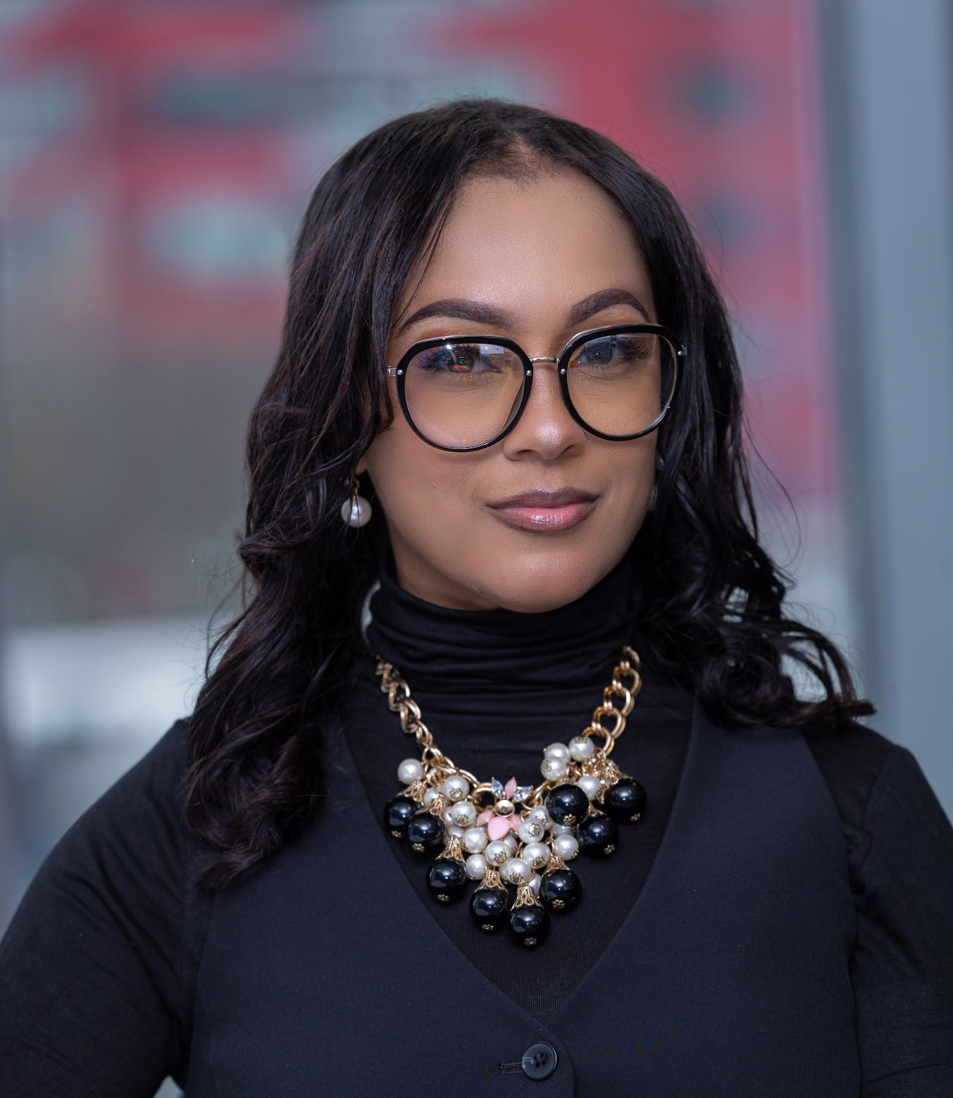 A woman wearing glasses and a necklace is standing in front of a red bus.