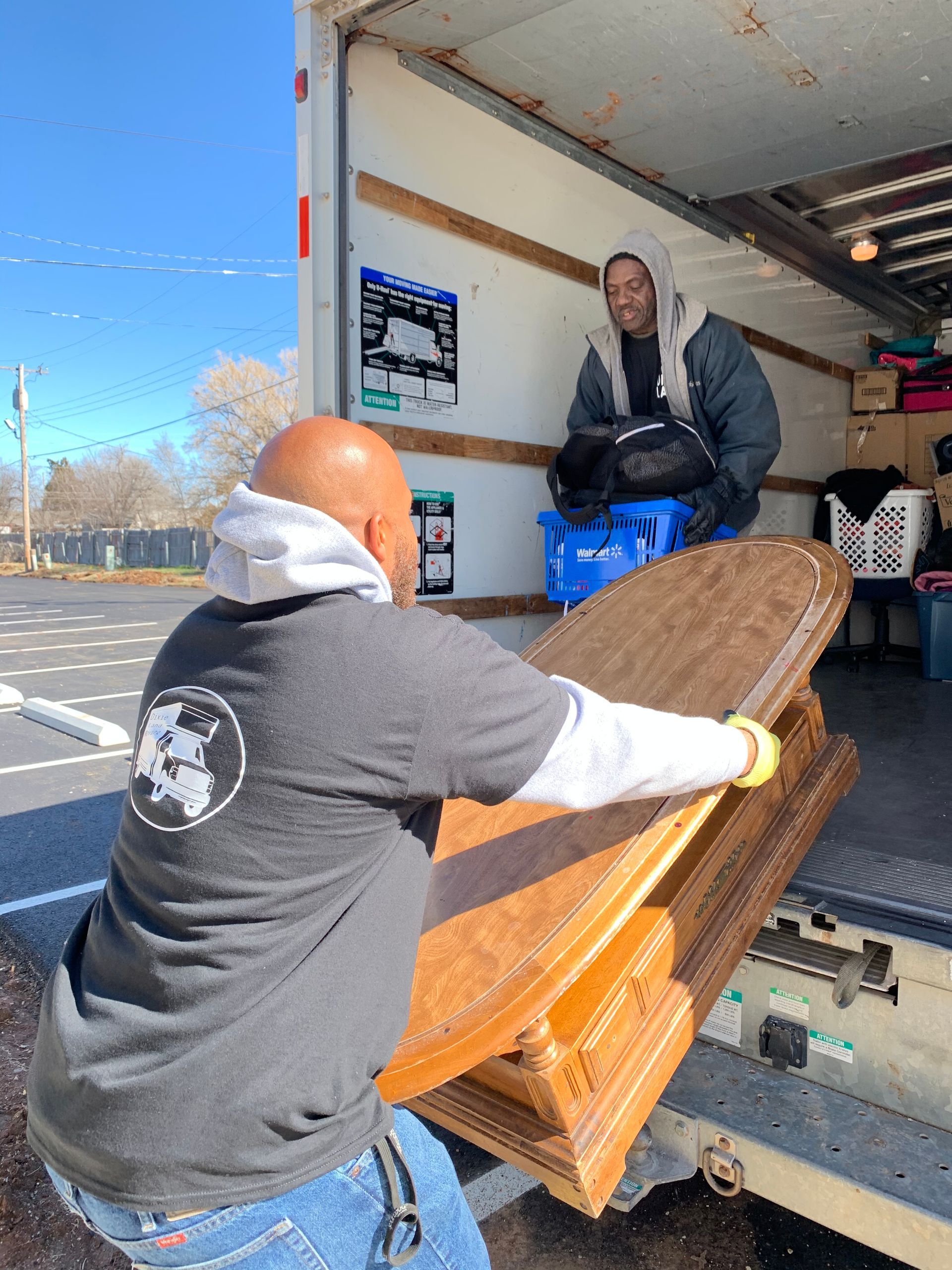 Two movers unload a large wooden tabletop from the back of a moving truck on a sunny day.
