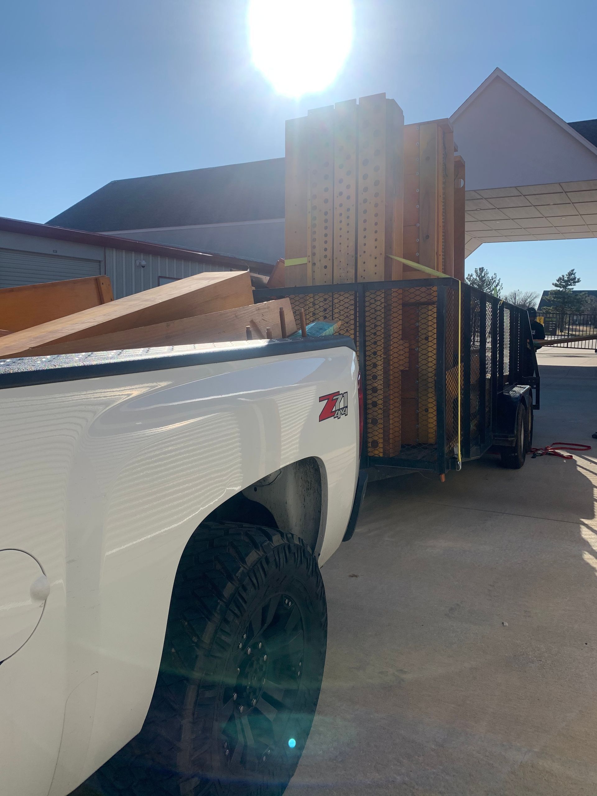 A white truck towing a trailer filled with large wooden structural beams parked under a canopy on a sunny day.