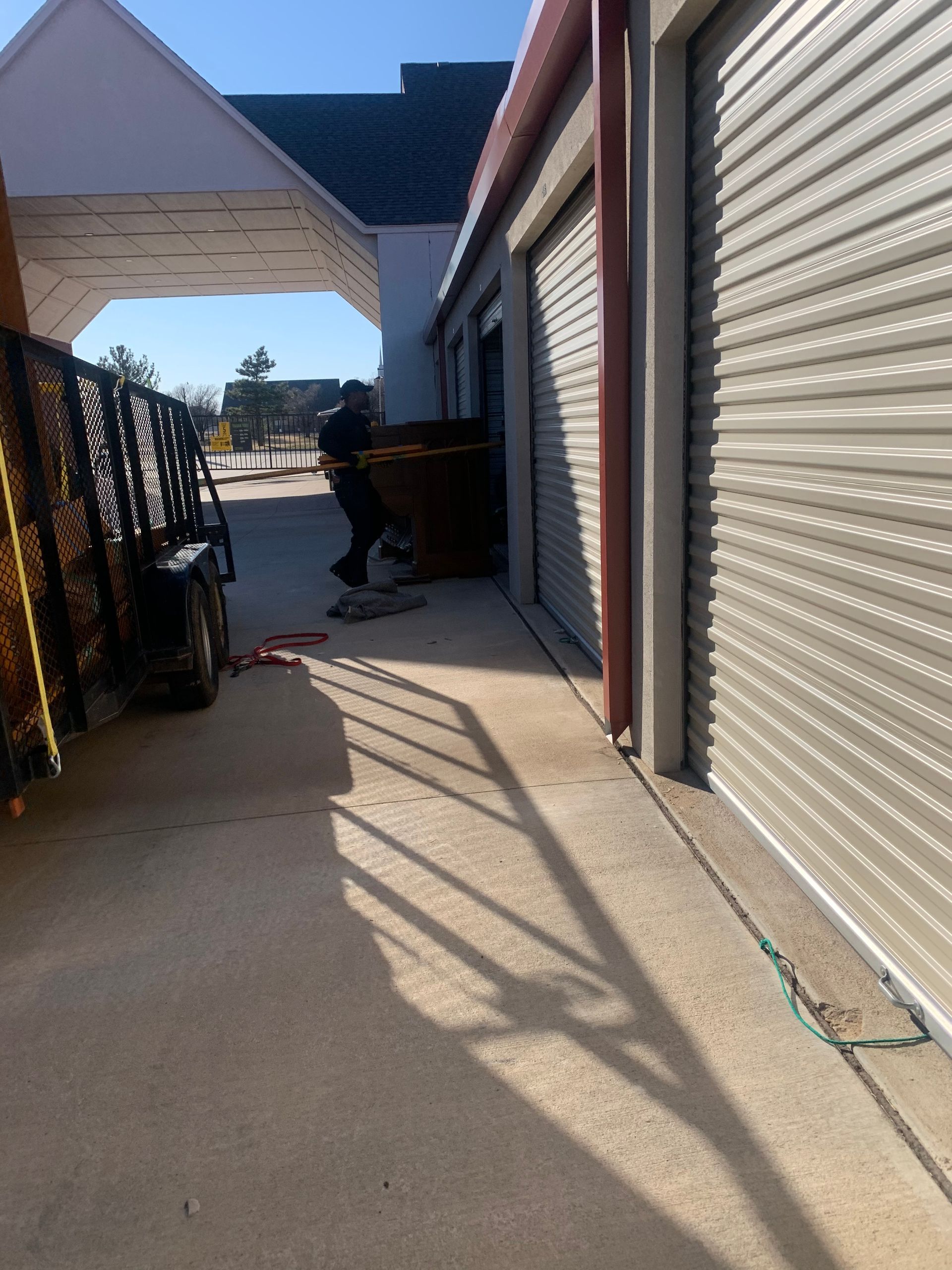 A person moves a large, dark box into a self-storage unit next to a trailer under a metal canopy.