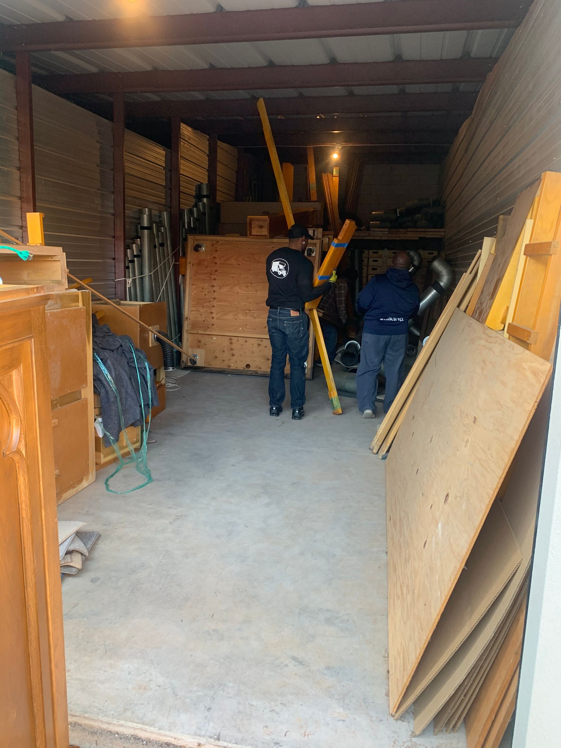 Two people move large wooden panels and planks inside a dimly lit storage unit.