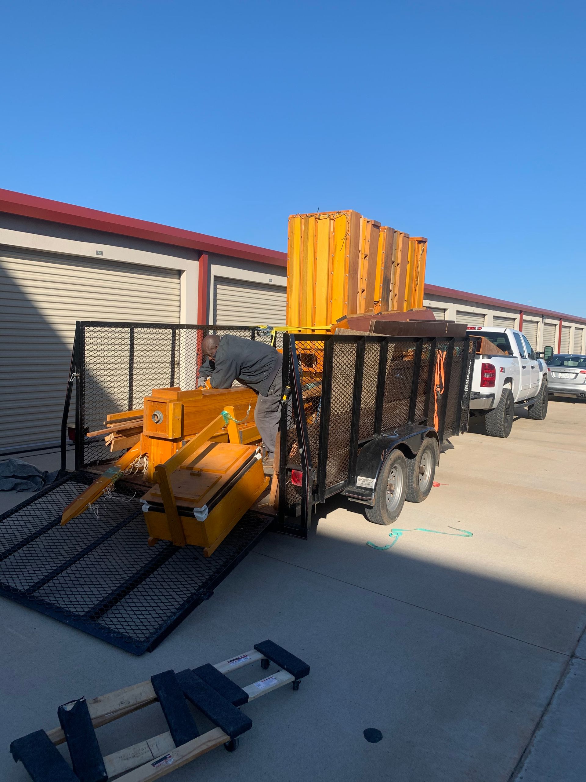 A person secures a large, yellow industrial log splitter onto a flatbed utility trailer parked in front of storage units.