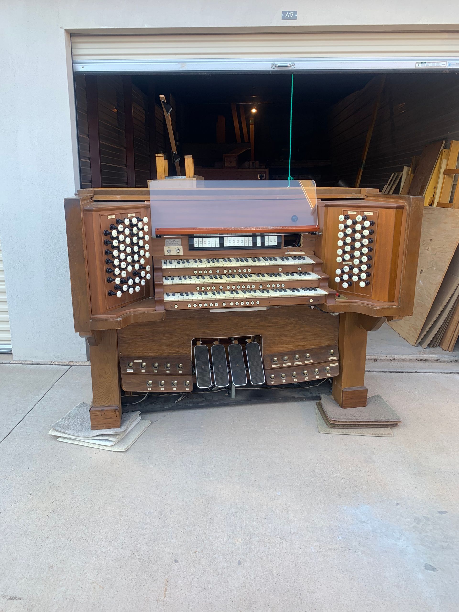 A three-manual wooden pipe organ console with many stops and pedals sits on a concrete driveway in front of a garage.