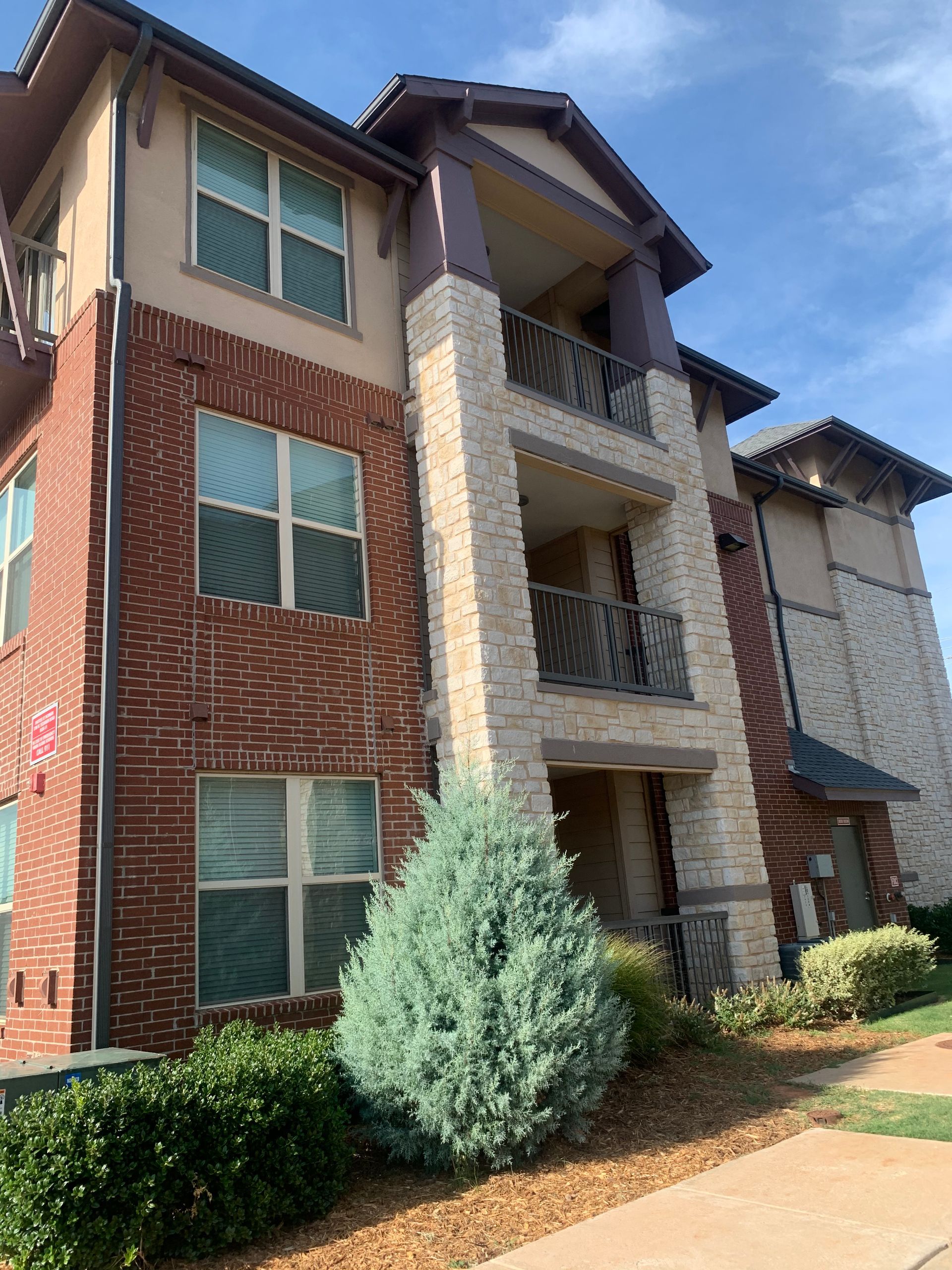 A three-story apartment building with red brick and light stone siding, featuring balconies and a bush in the foreground.