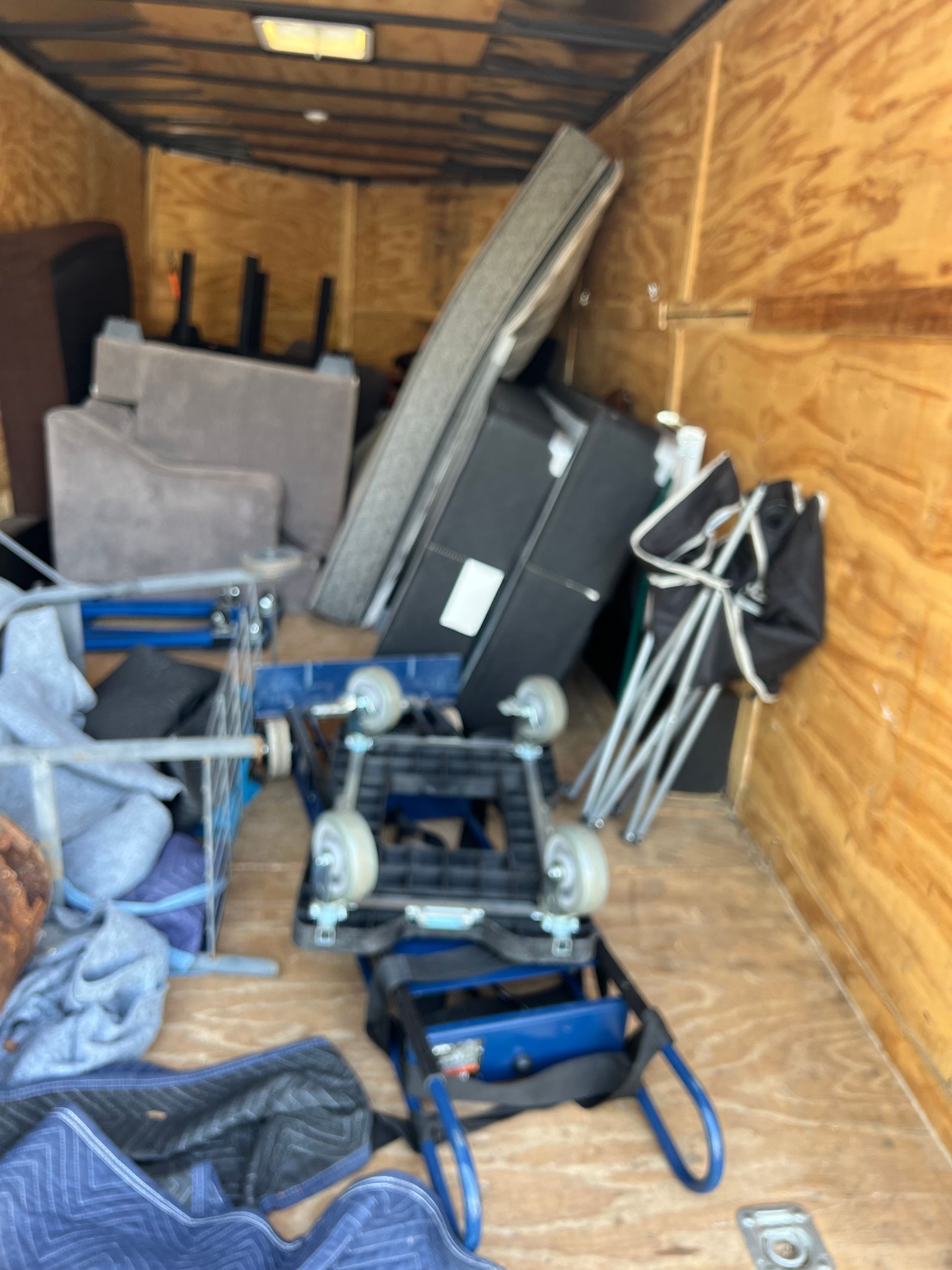 Interior of a wooden storage trailer containing a mattress, box spring, upholstered furniture, and moving dollies.