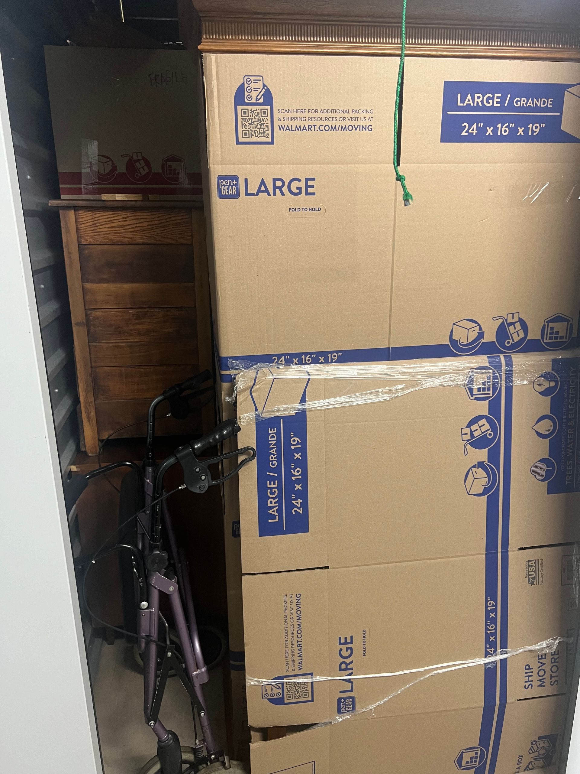 A tall stack of large cardboard moving boxes taped together, positioned in a tight storage space next to a purple walker.