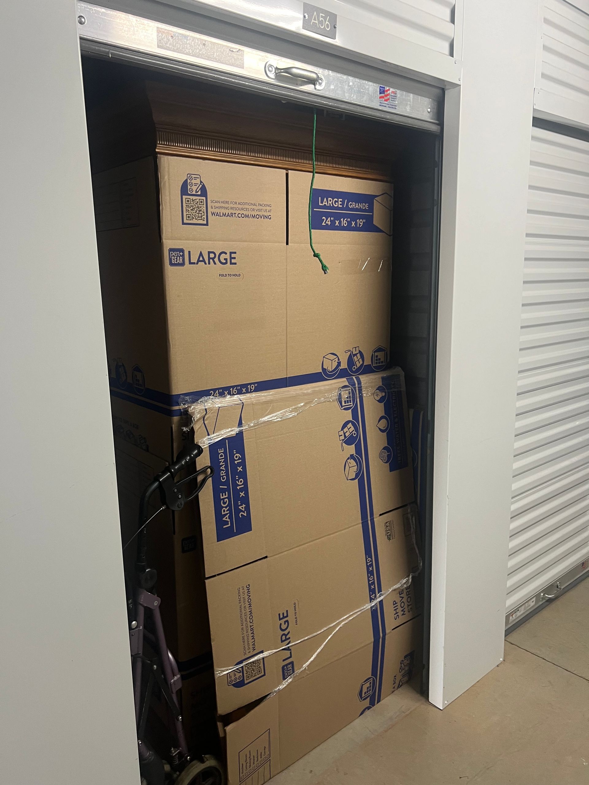 A storage unit filled with stacked large cardboard moving boxes and a metal hand truck parked on the left side.