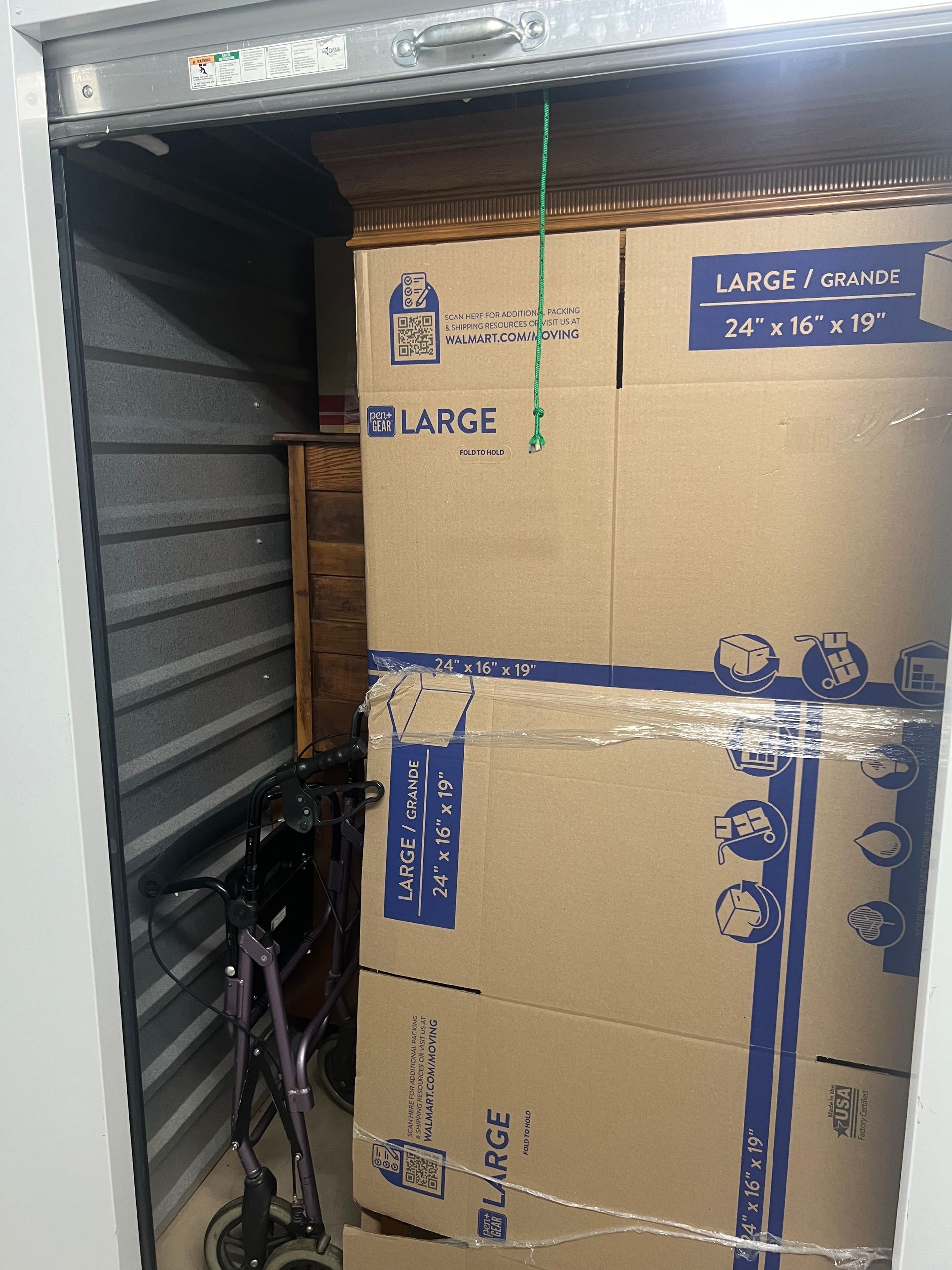 A stack of large, taped cardboard moving boxes fills most of a storage unit, with a walker visible to the left.