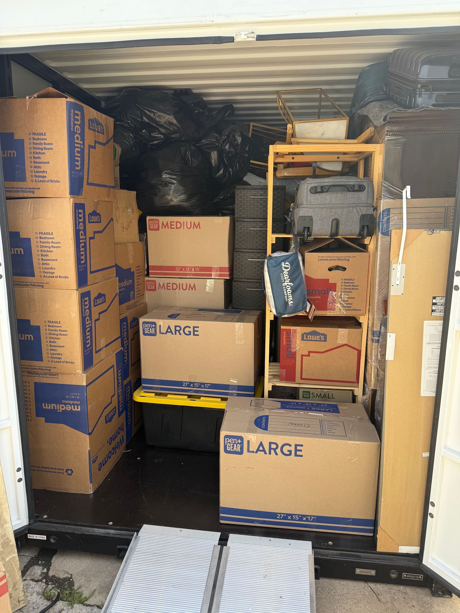 A storage unit filled with stacked cardboard moving boxes, black trash bags, and a wire shelving unit.