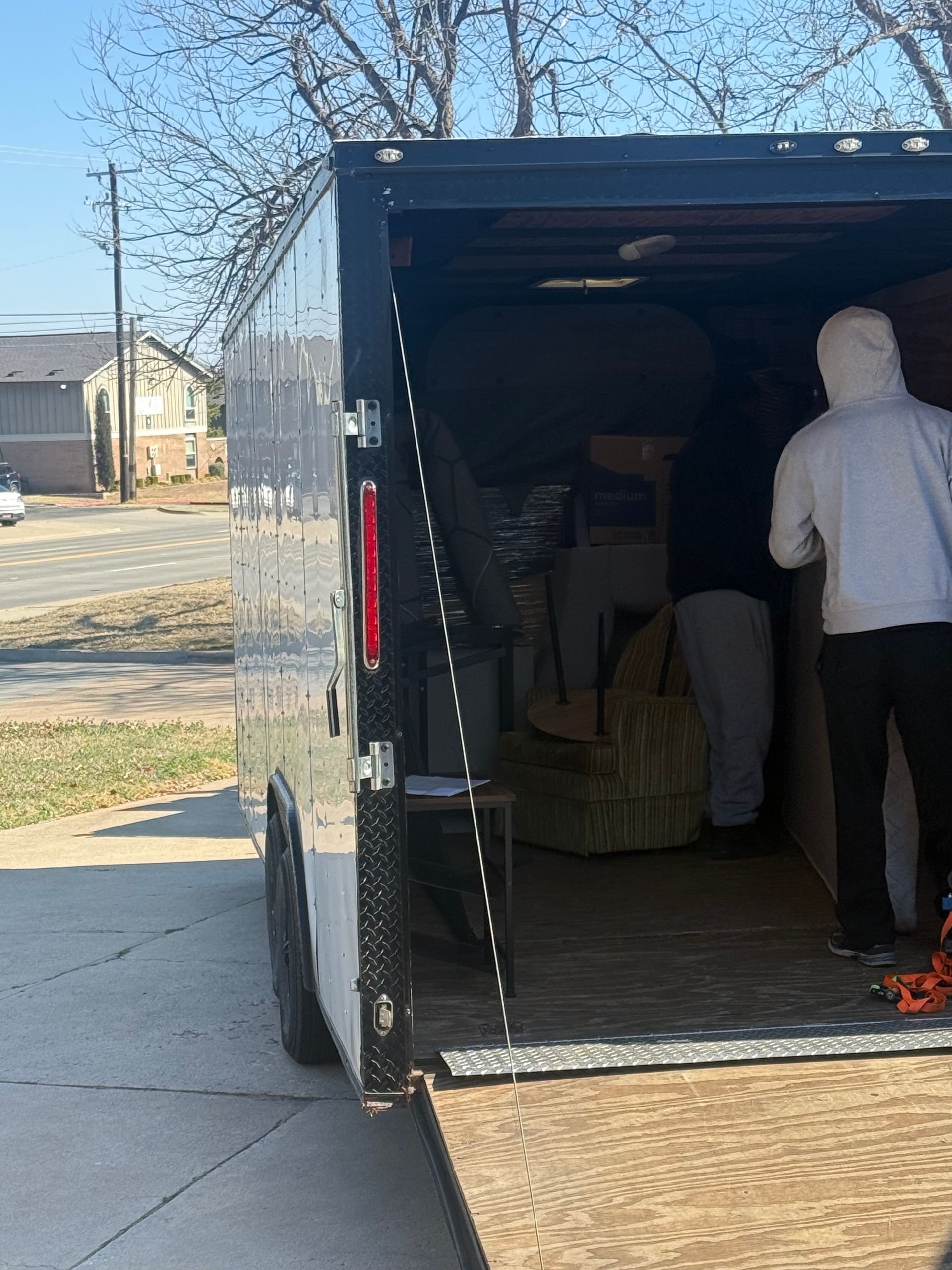 Two individuals move items into a black cargo trailer parked on a concrete driveway on a sunny day.