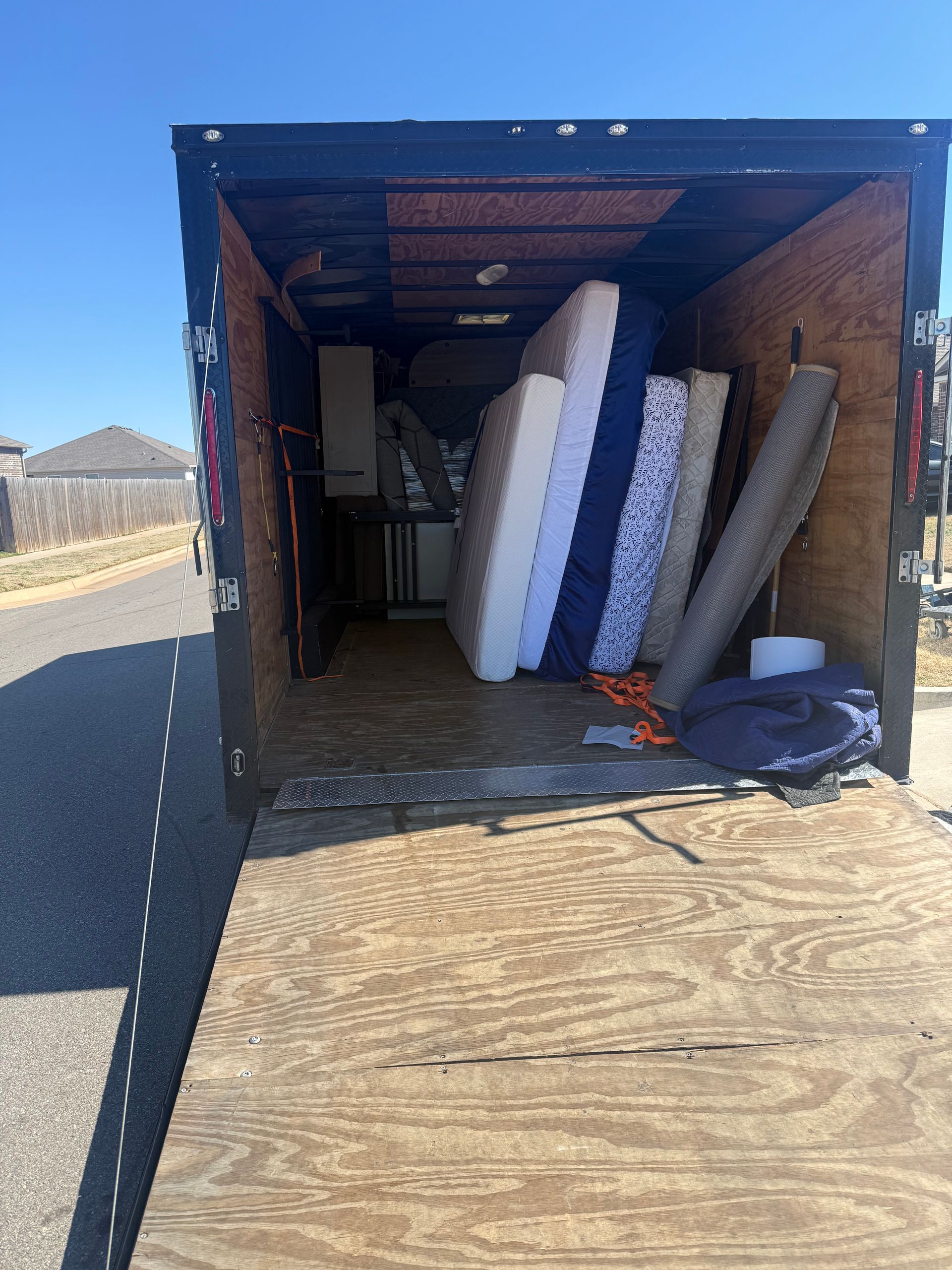 An open trailer parked on a street contains several upright mattresses of various patterns, colors, and sizes.