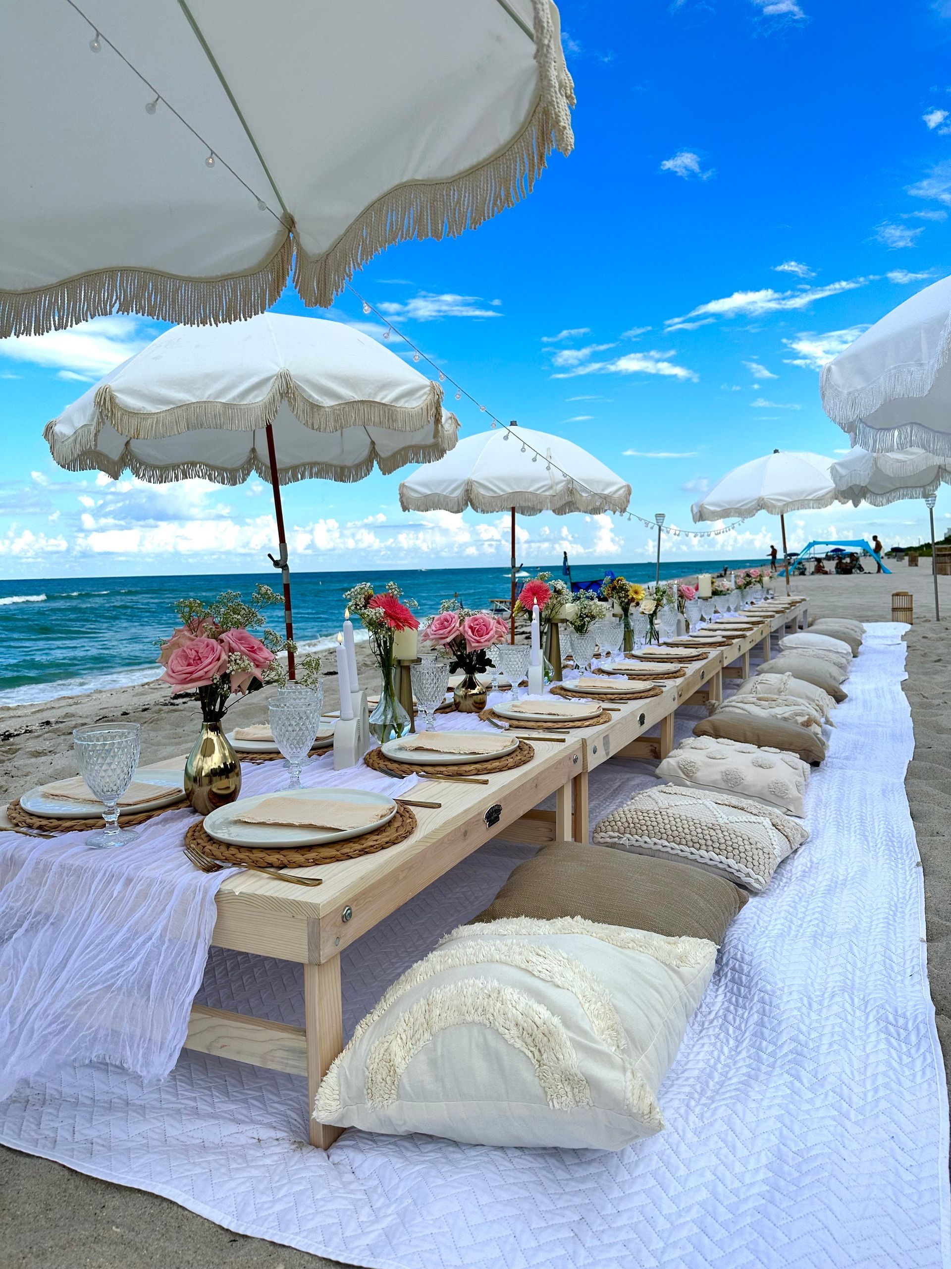 Beach picnic with long wooden table, cushions, and place settings. Blue and beige tones.