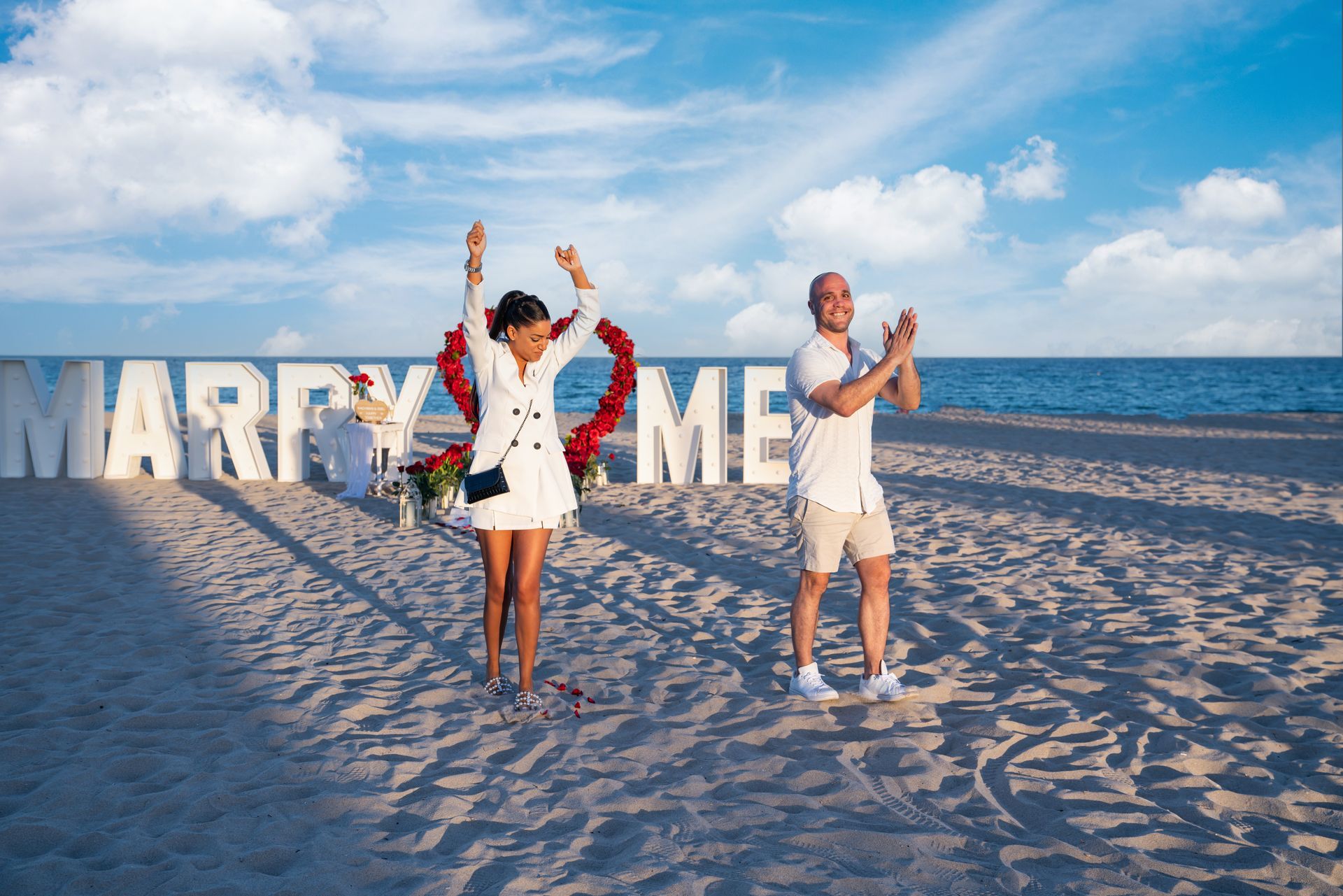 Woman raises arms, man claps, celebrating a beach proposal.