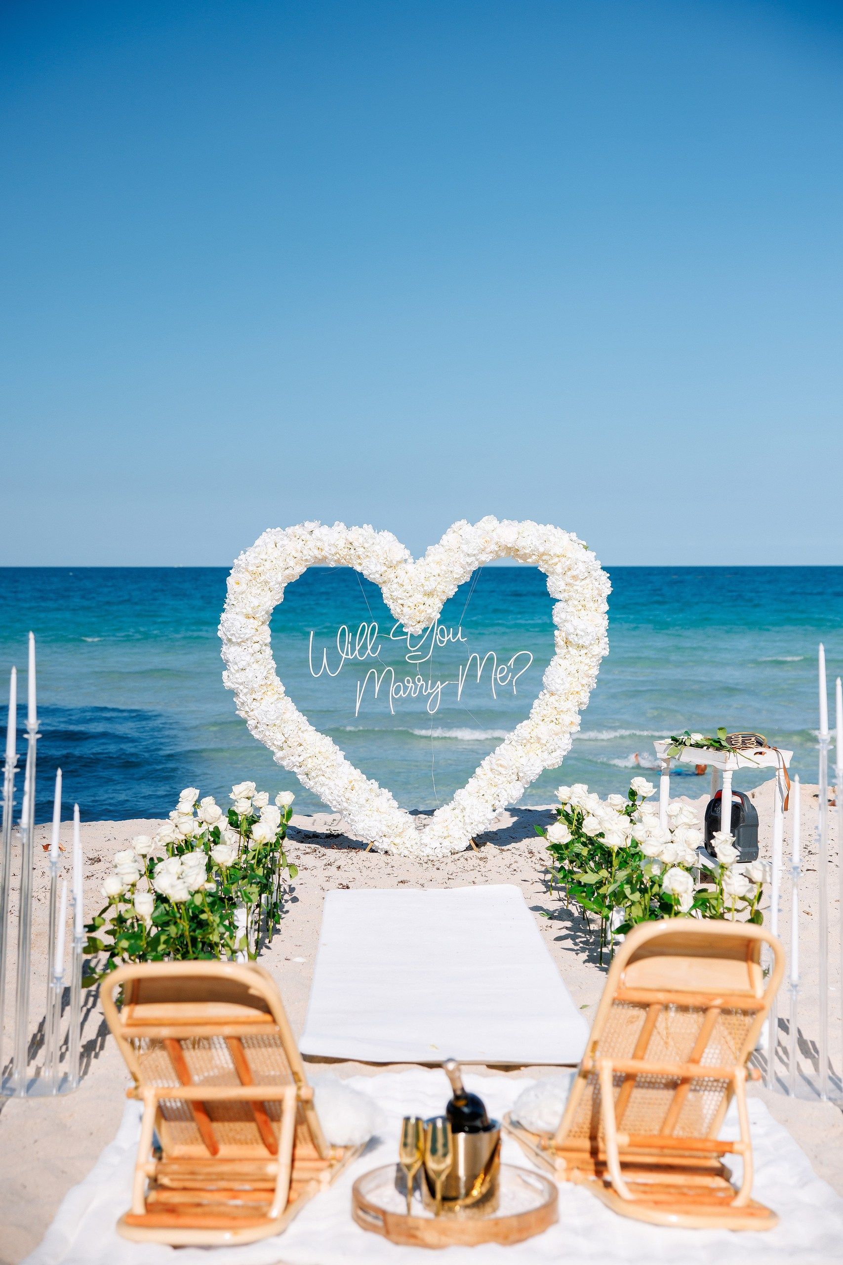 Beach wedding setup with heart-shaped floral arch. White flowers, ocean view, two chairs facing arch, sunny day.